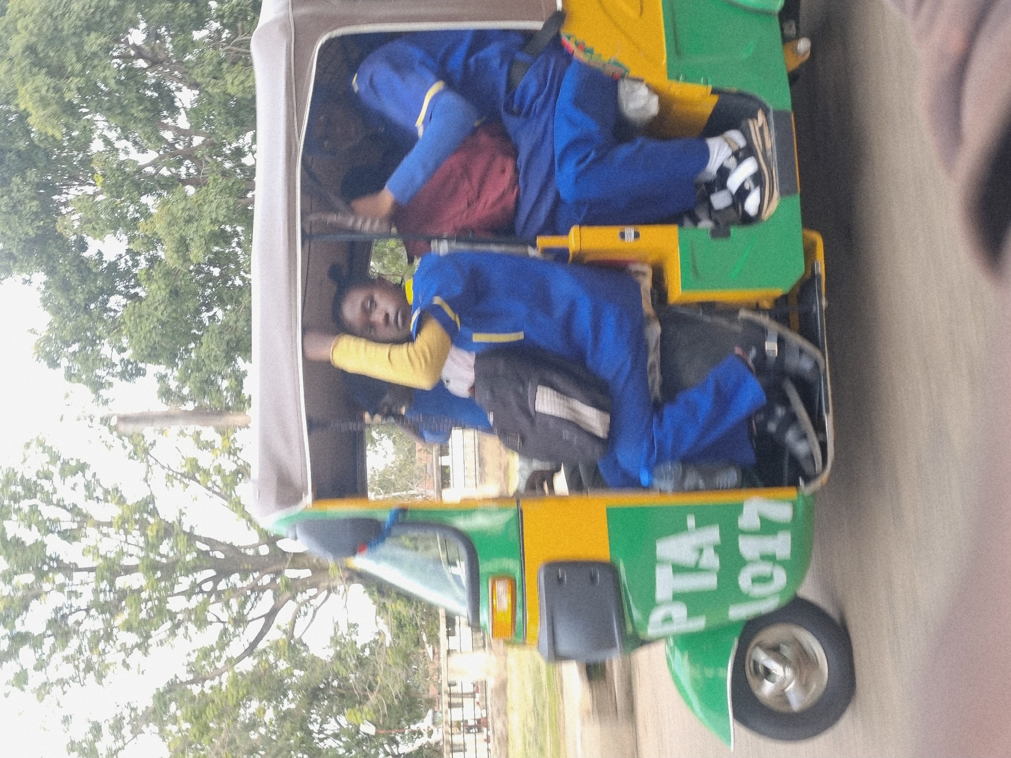 The image shows a bright, sunny day in Jos, Nigeria. The scene is dominated by a three-wheeled auto-rickshaw, commonly known as a "Keke Napep." The auto-rickshaw is painted green and yellow and has "PTA 1017" written on its side in white lettering.
Inside the auto-rickshaw, two individuals are visible. One person is lying down, wearing a blue uniform with yellow trim and white socks. Another person, also in a similar blue uniform with yellow trim and a yellow shirt underneath, is lying in the lower compartment of the vehicle. The body language suggests they might be resting or traveling long distances.
The auto-rickshaw is likely on a road, with a tree and foliage visible in the background, indicating an outdoor setting with some greenery. The overall impression is one of everyday life and transportation in Nigeria, with the bright colors and casual arrangement of people within the vehicle being characteristic of the region. The orientation of the photo is somewhat tilted, as if taken from a moving vehicle or by someone holding the camera at an angle.