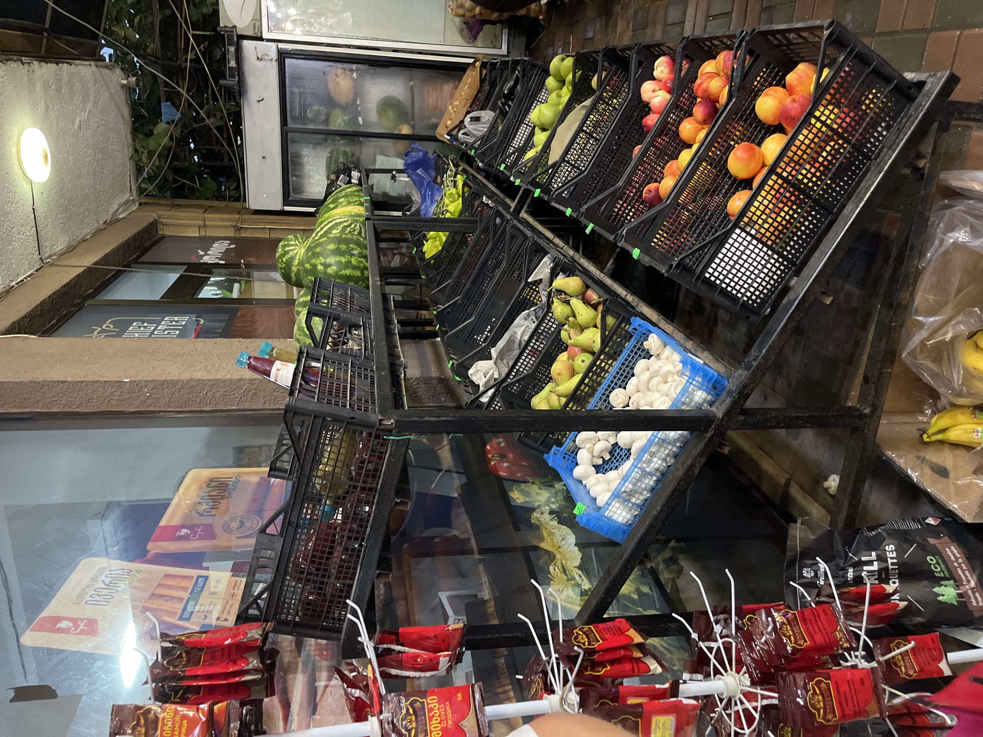 The image depicts an outdoor fruit and vegetable stall located in the city of Ureki, Georgia. The stall features a metal shelving unit displaying a variety of produce in black plastic crates. On the upper shelves, watermelons are prominently featured, alongside other fruits like pears and peaches on lower shelves. There are also crates containing mushrooms and what appear to be yellow bell peppers. Packaged goods, including bags of spices and briquettes, are visible on the left side of the image. In the background, a refrigerated display case is partially visible, suggesting it's part of a larger shop. The environment appears to be a street-level establishment, with some foliage and a concrete or tiled floor visible. No people are directly interacting with the produce or the stall in a significant way. The lighting suggests it is daytime, with some artificial light sources visible overhead.