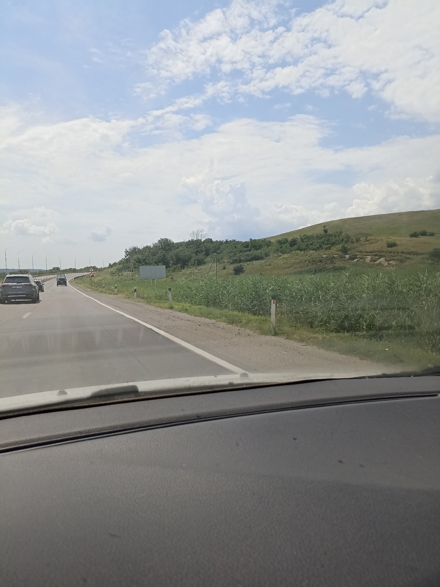 The image shows a view from inside a car, looking out at a highway on a cloudy day. The road curves to the left, with several cars visible in the distance. To the right of the road, there is a grassy hill covered in trees and vegetation. The sky is filled with white, fluffy clouds against a light blue background. The foreground of the image shows the dashboard of the car. The scene is a typical highway landscape, likely in or around Mineralnye Vody, Russia, given the vegetation and road infrastructure.