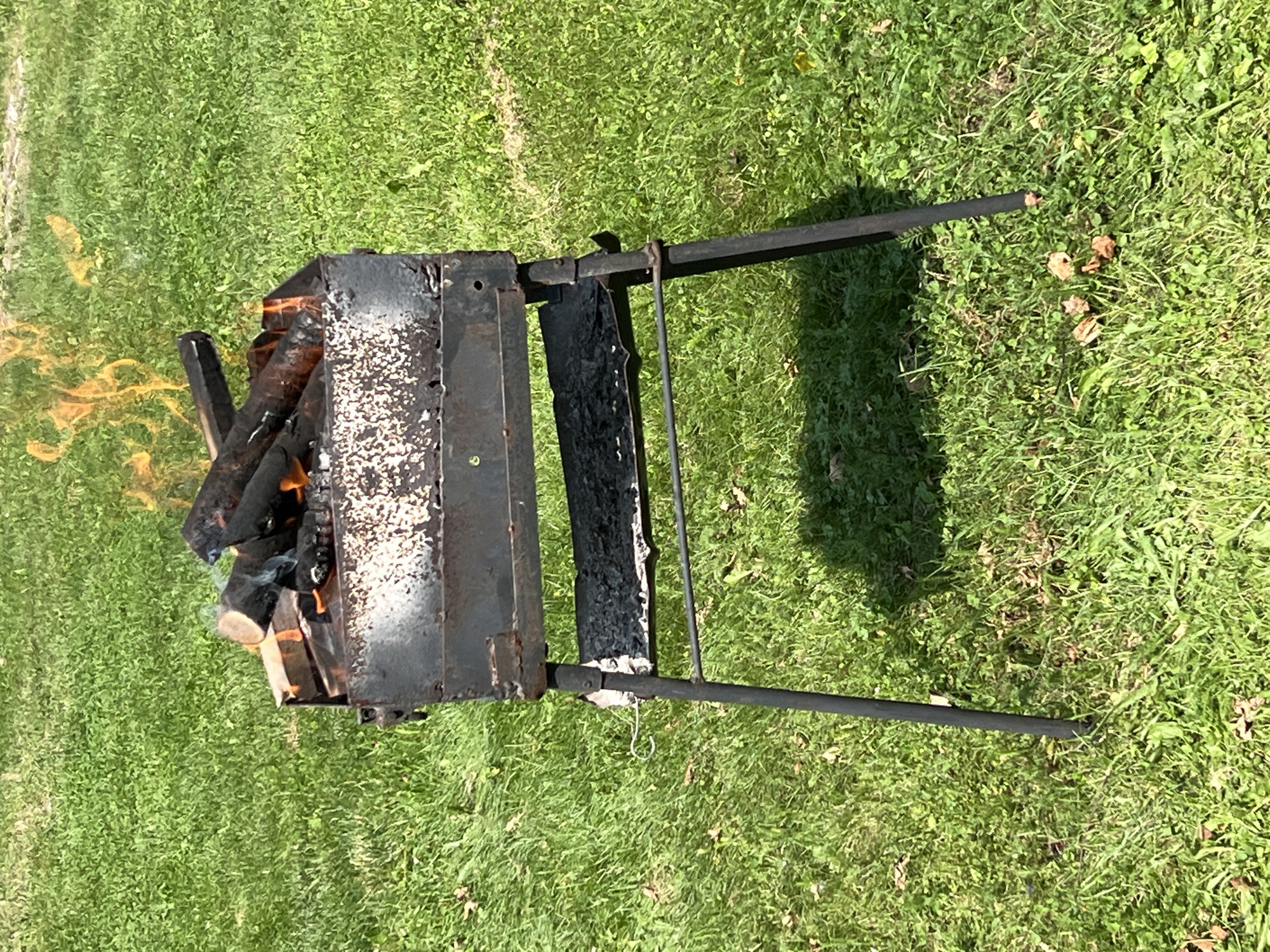 A black metal barbecue grill with burning wood is situated on a green grassy lawn. The grill is raised on four black legs and has a lower rack containing ash. Flames are visible in the upper portion of the grill, consuming the logs. The background consists of a dense green lawn with some scattered brown leaves. The scene is outdoors in daylight.