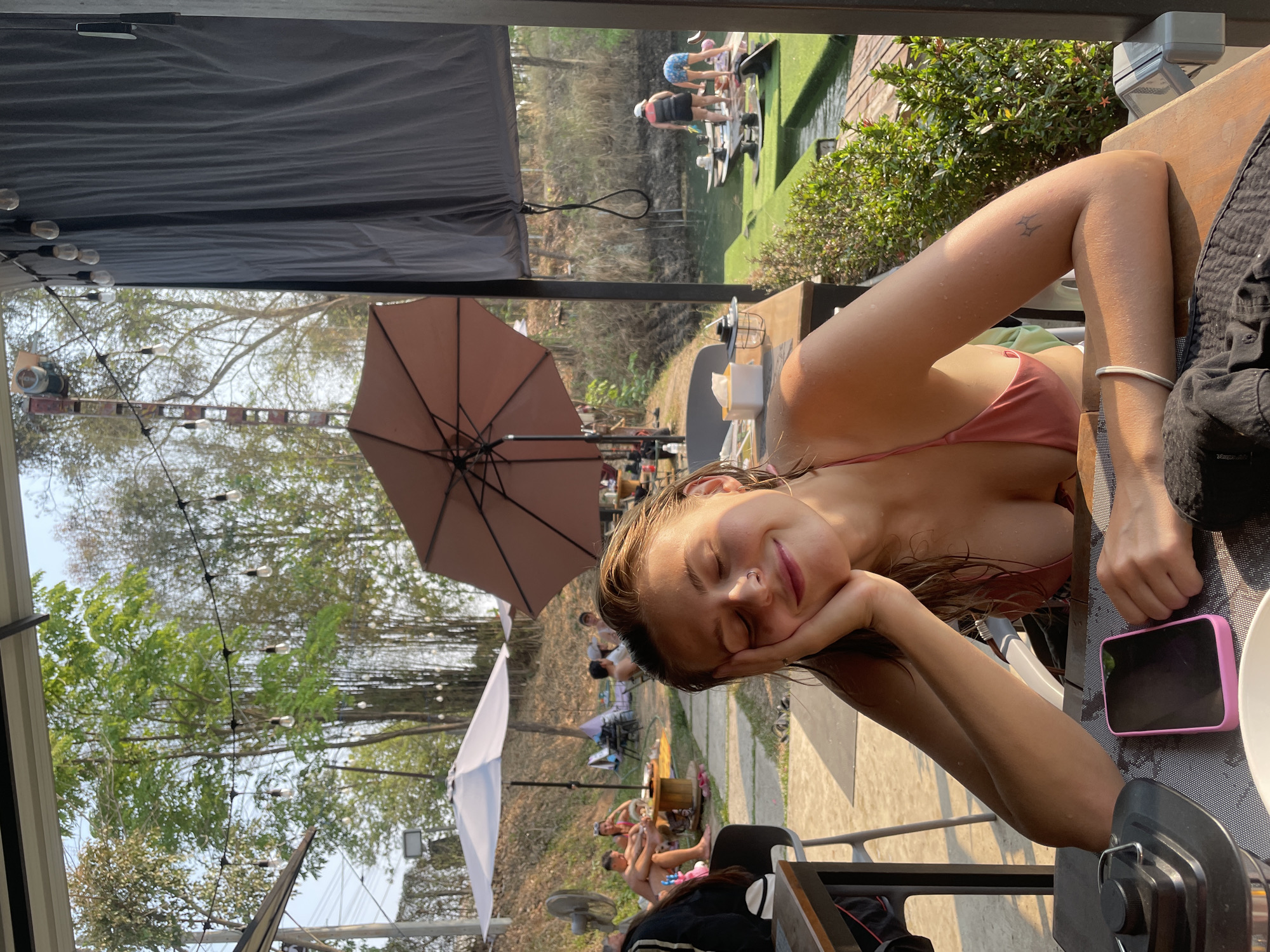 A young woman is reclining at a table in an outdoor setting at a location in Tambon Nam Phrae, Thailand. She is wearing a swimsuit and resting her head on her hand. A mobile phone is lying next to her on the table. An outdoor seating area with umbrellas and other patrons can be seen in the background. A body of water with people participating in water activities is also visible in the distance.