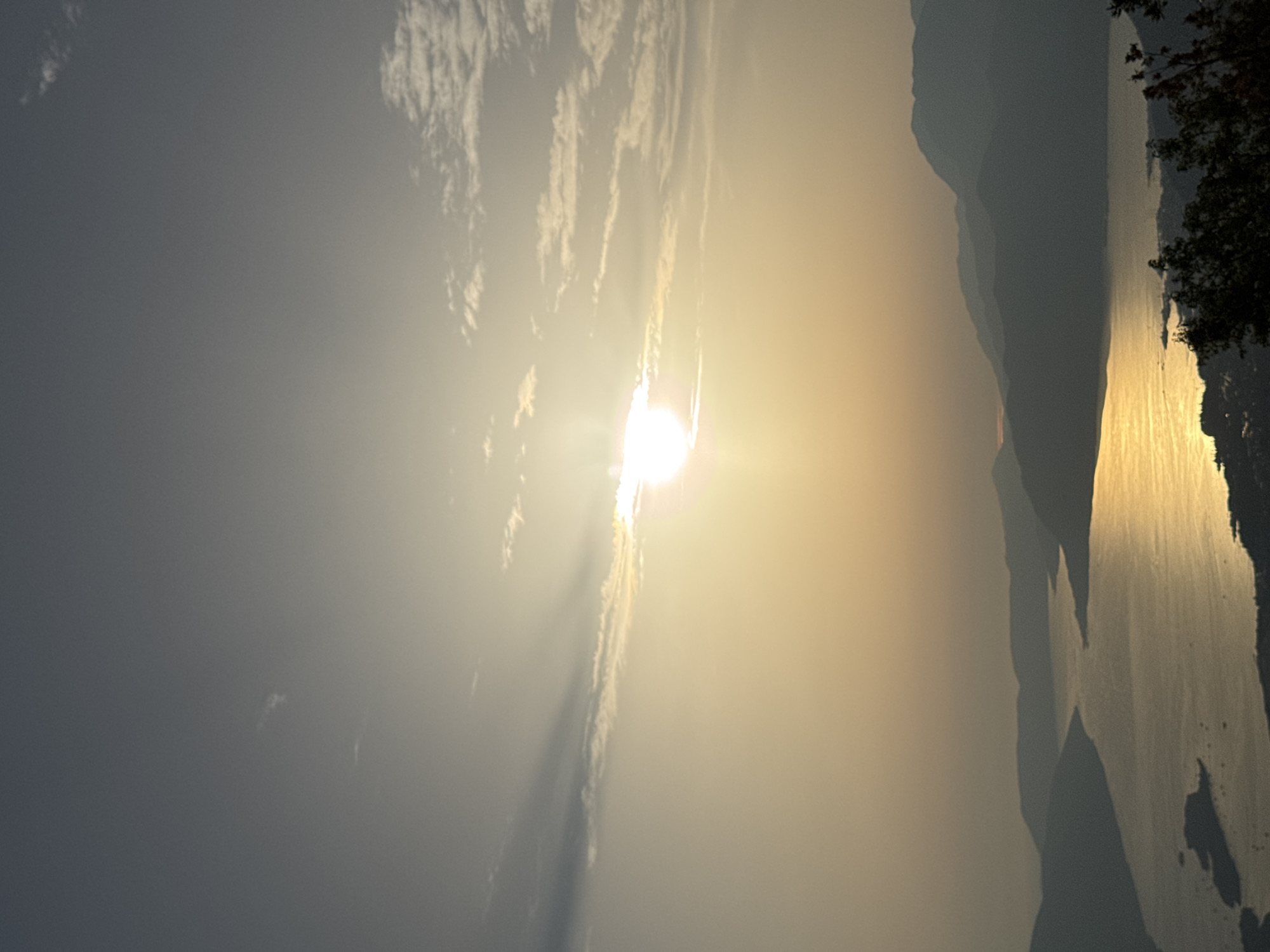 The image shows a serene sunset over a bay in Špiljari, Montenegro. The sun is partially obscured by a thin cloud, casting a warm, golden glow across the water. The calm sea reflects the sunlight, creating a shimmering path. Silhouetted mountains surround the bay, their peaks fading into the hazy sky. The foreground features a hint of lush green trees, adding a touch of nature's presence to the tranquil scene. No people or specific objects are clearly discernible, emphasizing the natural beauty of the landscape. The overall impression is one of peace and quiet, a typical evening vista in a coastal Montenegrin town.