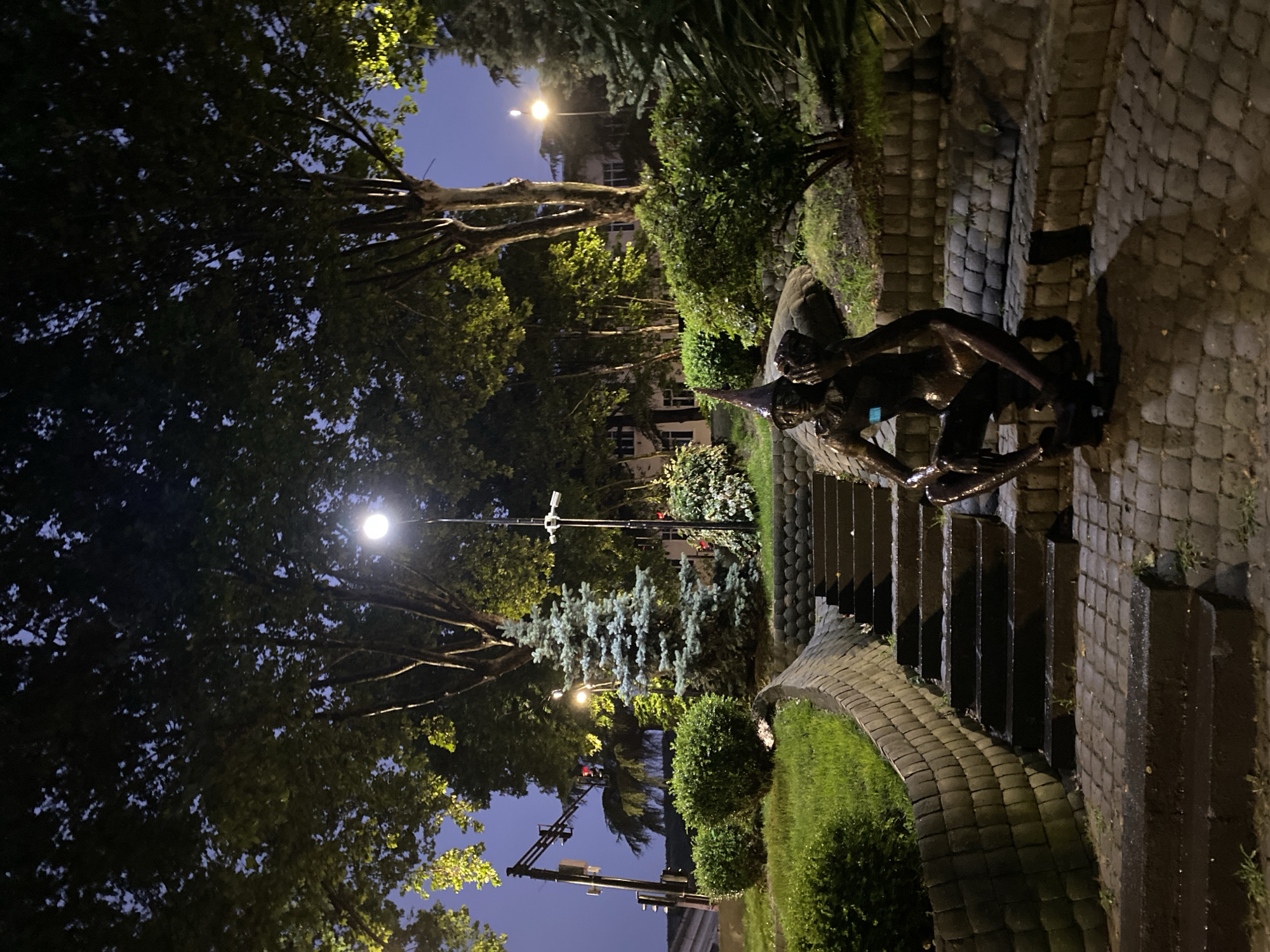 The image depicts a nighttime scene with a statue of a figure wearing a conical hat, sitting on a stone base. The statue is located near a set of stone stairs leading up a grassy embankment. Surrounding the area are trees, bushes, and streetlights illuminating the scene. In the background, buildings with illuminated windows are visible. A traffic light is also present on the left side of the image. The overall ambiance suggests an outdoor public space, possibly a park or a plaza, in Tbilisi, Georgia.