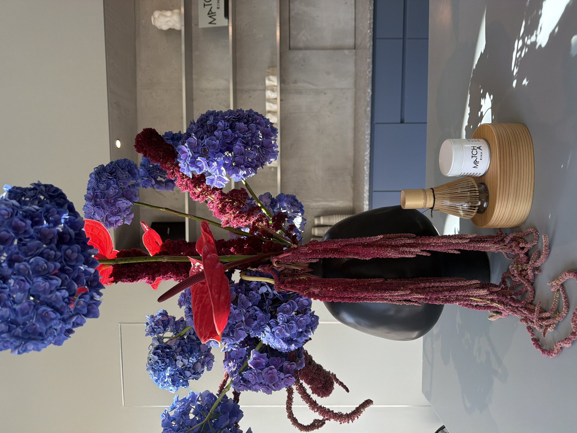 The image depicts a still life arrangement on a grey table, likely in a modern kitchen or dining area. In the foreground, a large, dark, bulbous vase holds a dramatic floral arrangement. The flowers are a mix of vibrant blue hydrangeas, striking red anthuriums, and cascading, deep red amaranth. To the right of the vase, a bamboo matcha whisk sits on a light wood circular base. Next to the whisk is a small white jar with a label that reads "MATCHA RINA". In the background, there are light grey shelves mounted on a textured wall. The shelves hold a few small decorative items, including a white bust on the top shelf and some stacked rectangular objects on the middle shelf. Below the shelves are blue kitchen cabinets. The overall impression is one of contemporary elegance and design, with a focus on natural elements and a touch of Japanese aesthetic represented by the matcha set. There are no people in the image. The lighting suggests an indoor setting, possibly with natural light filtering in from a window, casting shadows on the table. It is not possible to determine the specific location in Munich, Germany from the image alone.
