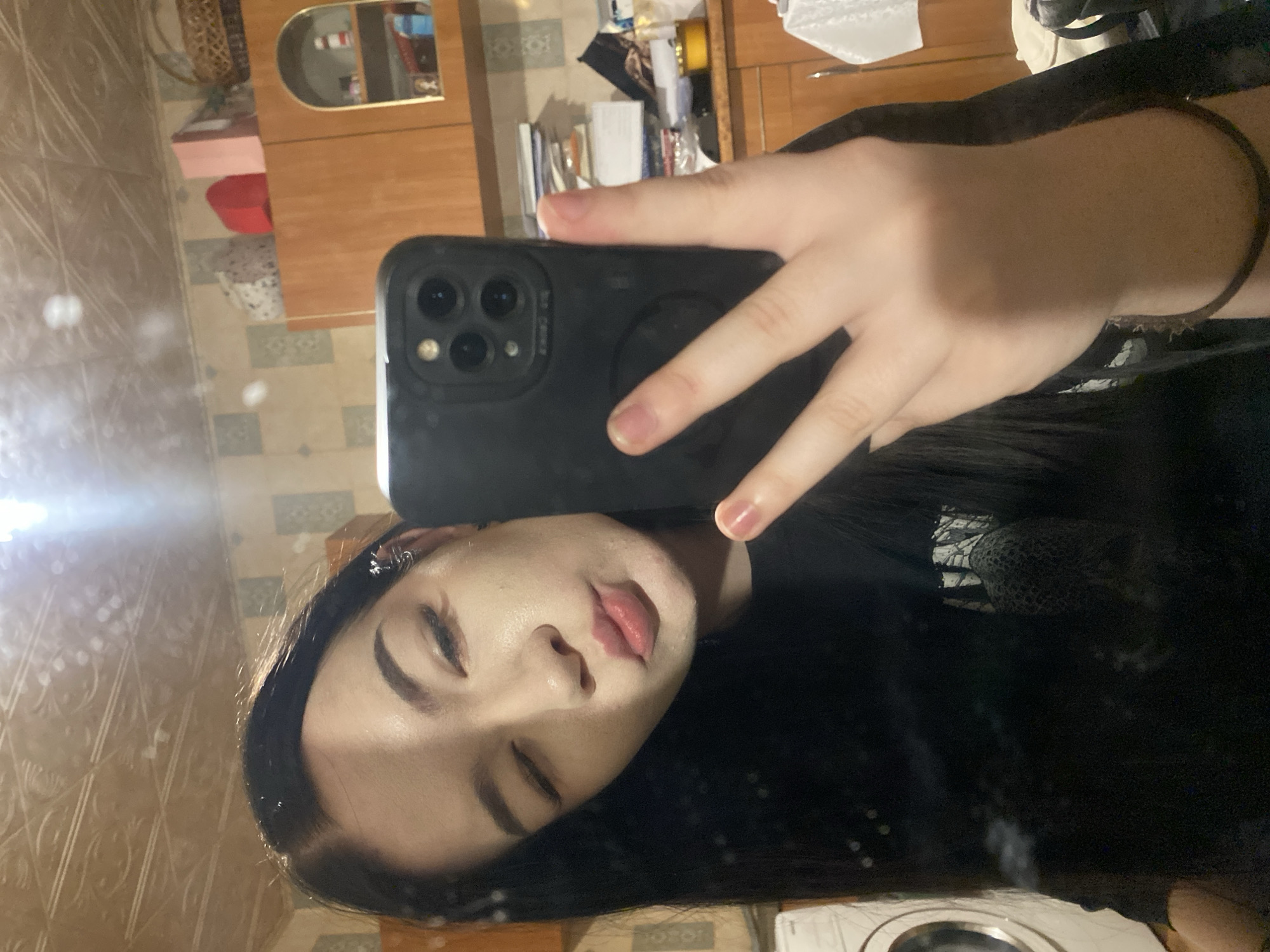 A person with closed eyes is taking a selfie in a mirror. The person has black hair and is wearing a black shirt. The reflection in the mirror shows a room with tiled walls and a wooden cabinet. The phone is a black iPhone with three lenses on the back. It is not possible to determine the exact location in Ternopil, Ukraine.