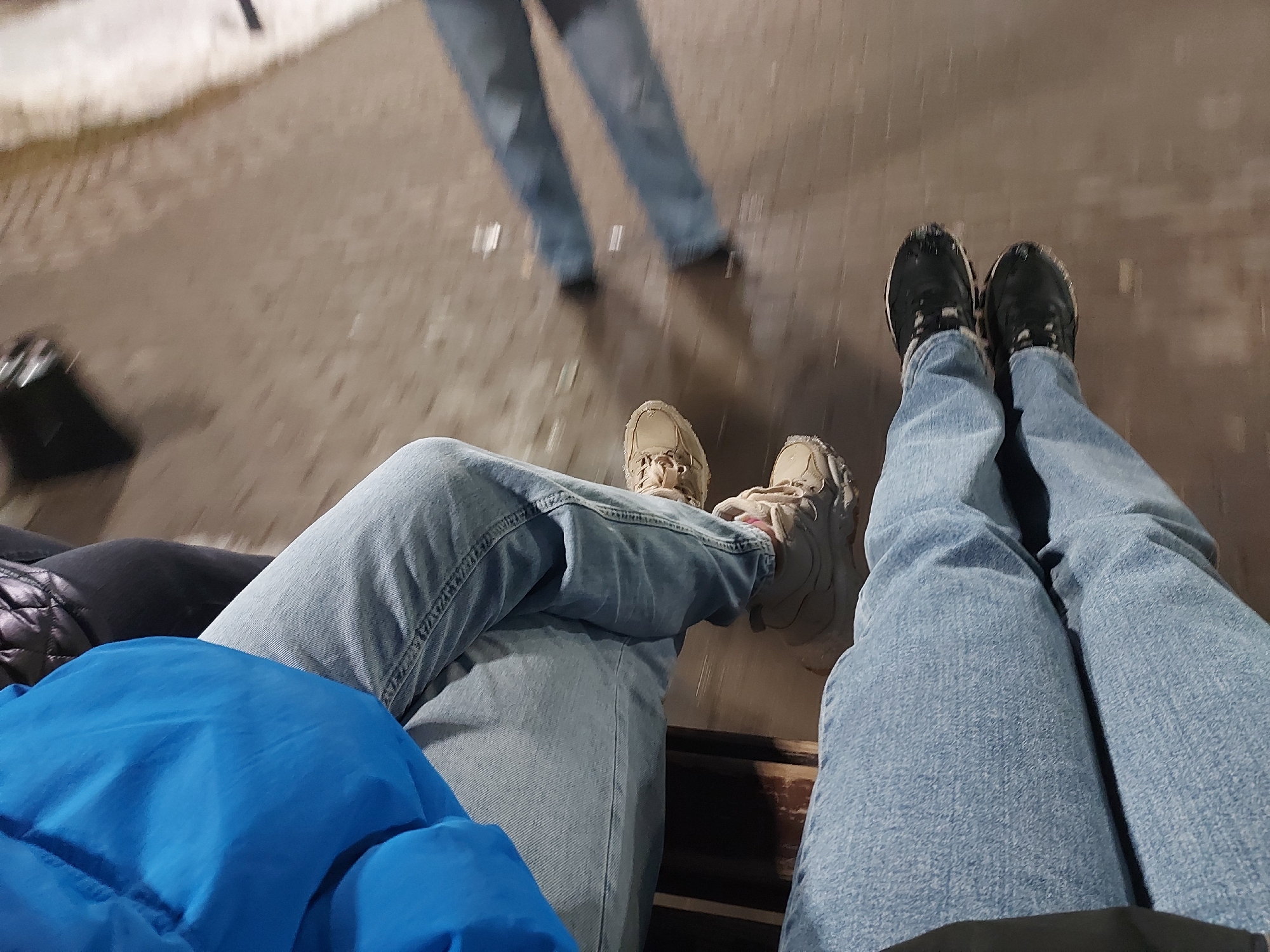 The image shows three pairs of legs and shoes. Two pairs are sitting side-by-side on a bench, wearing light blue jeans. One pair has beige sneakers with pink laces, and the other has black sneakers. A third pair of legs, wearing blue jeans, is standing a short distance away. The ground appears to be a paved surface, possibly wet. The background is blurry and out of focus, but suggests an outdoor setting, possibly a street or plaza. It is difficult to confirm the location as Chaikovsky, Russia, without additional context.