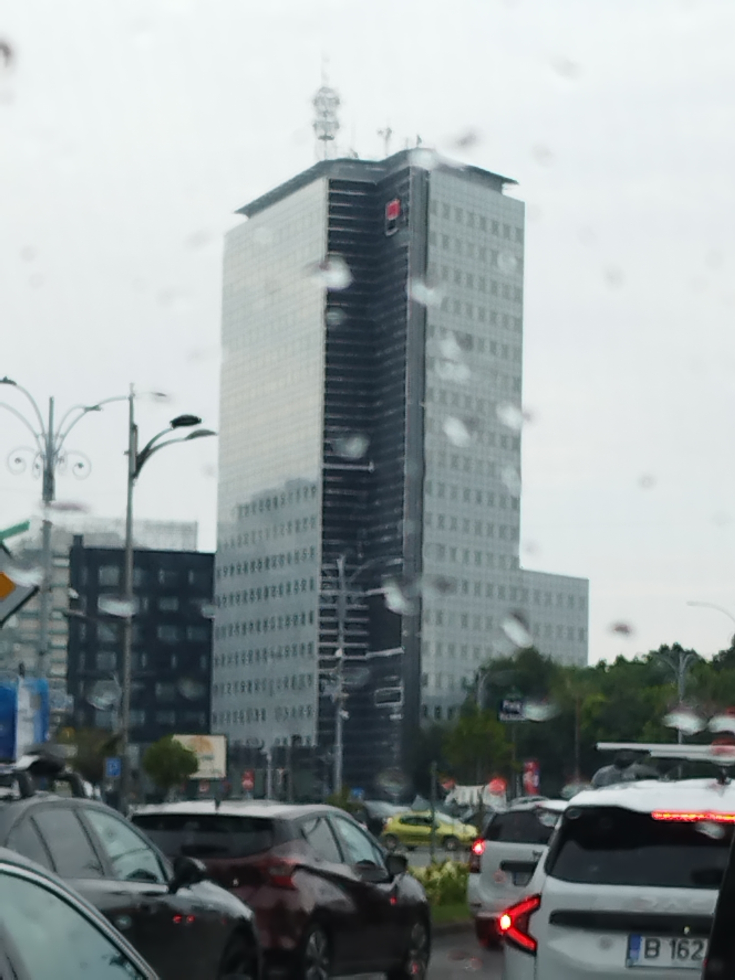 The image captures a street-level view in Bucharest, Romania, on a rainy day, evidenced by water droplets on the camera lens and a hazy atmosphere. In the background, a prominent modern skyscraper, partially under construction or renovation with visible scaffolding, dominates the skyline. The building features a glass facade and appears to be a significant commercial or office building. In the foreground and midground, a traffic jam is evident, with several cars of various makes and models visible. The vehicles are closely packed, suggesting slow-moving or stopped traffic. The street is paved, and there are indications of urban infrastructure, including streetlights and traffic signs. Trees and other buildings are also visible, contributing to the urban landscape. The overall impression is one of a busy city street experiencing a common urban phenomenon on a wet day.