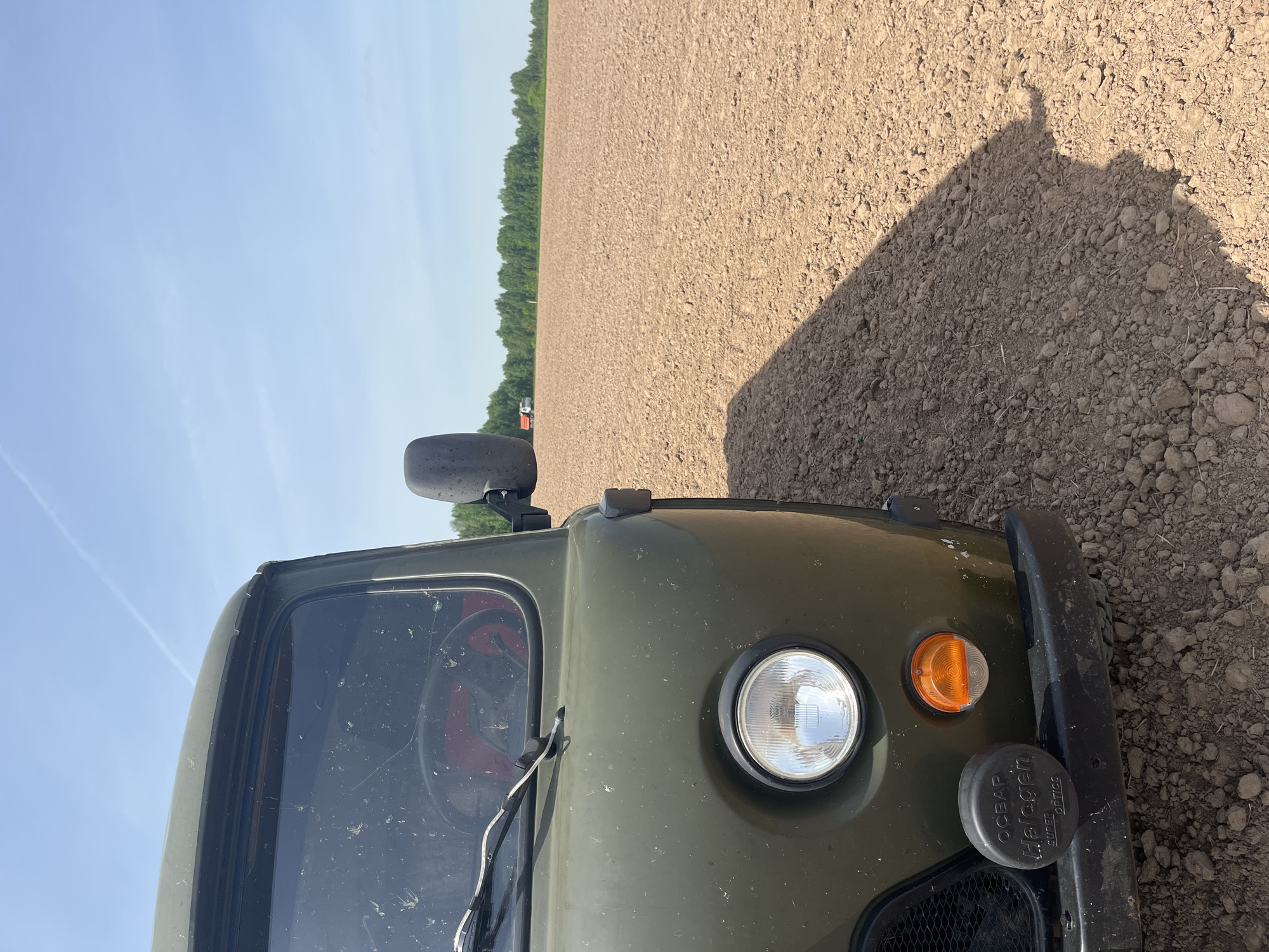 A close-up, angled shot shows the front of an olive-green vehicle parked on a tilled field. The vehicle's headlight, turn signal, bumper, and a portion of the windshield are visible. The field stretches out behind the vehicle towards a line of trees in the distance under a blue sky. The vehicle casts a shadow onto the ground. The location and city are unknown.