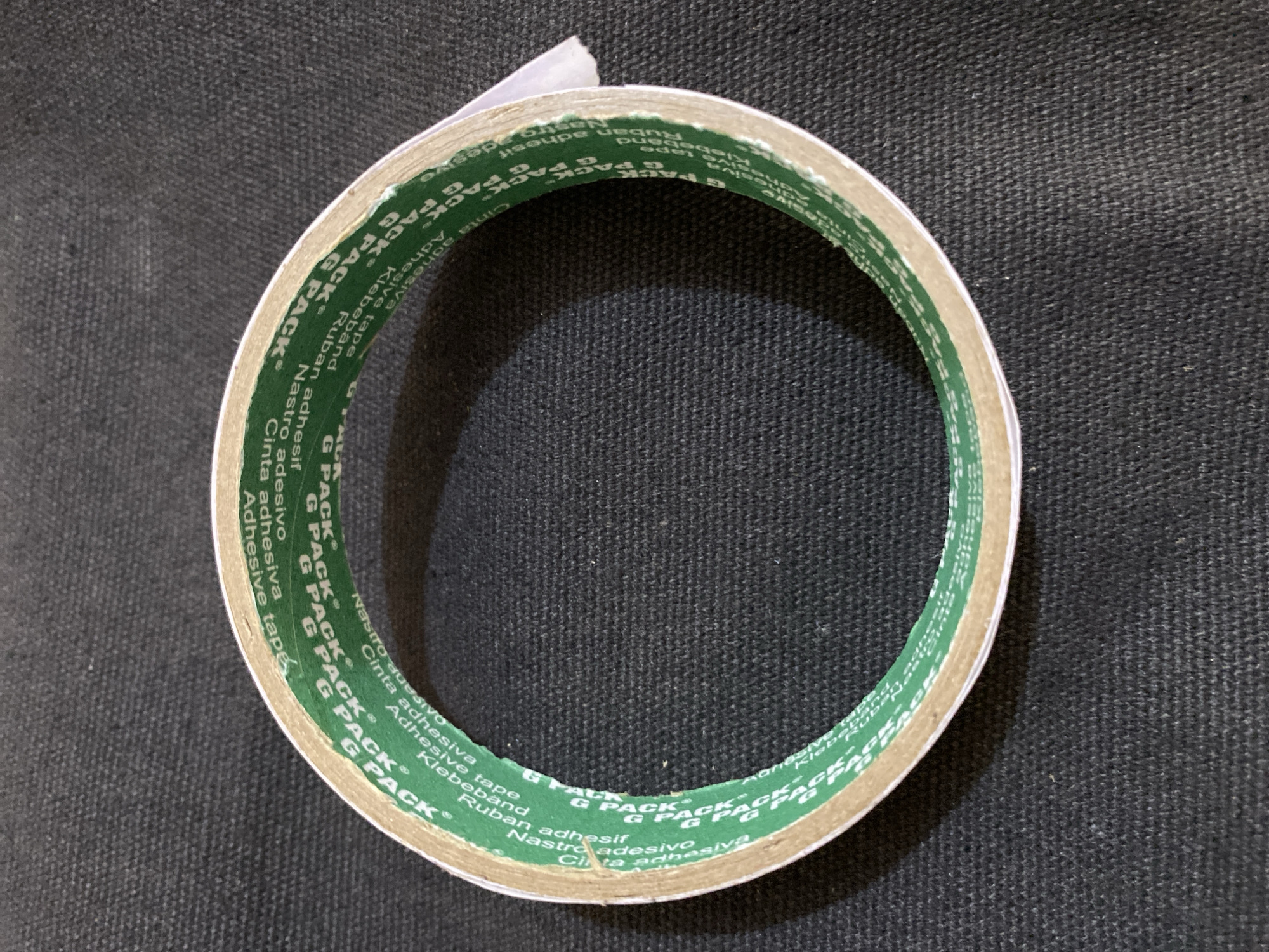 A roll of double-sided adhesive tape is centered on a dark, textured surface, likely a fabric. The tape is green with repeated white text reading "G PACK®" and "Cinta Adhesiva Tape" along the edge. The tape has a beige backing paper peeking out from one side. The lighting casts a subtle shadow around the tape. There is no indication of a city or location within the image itself.