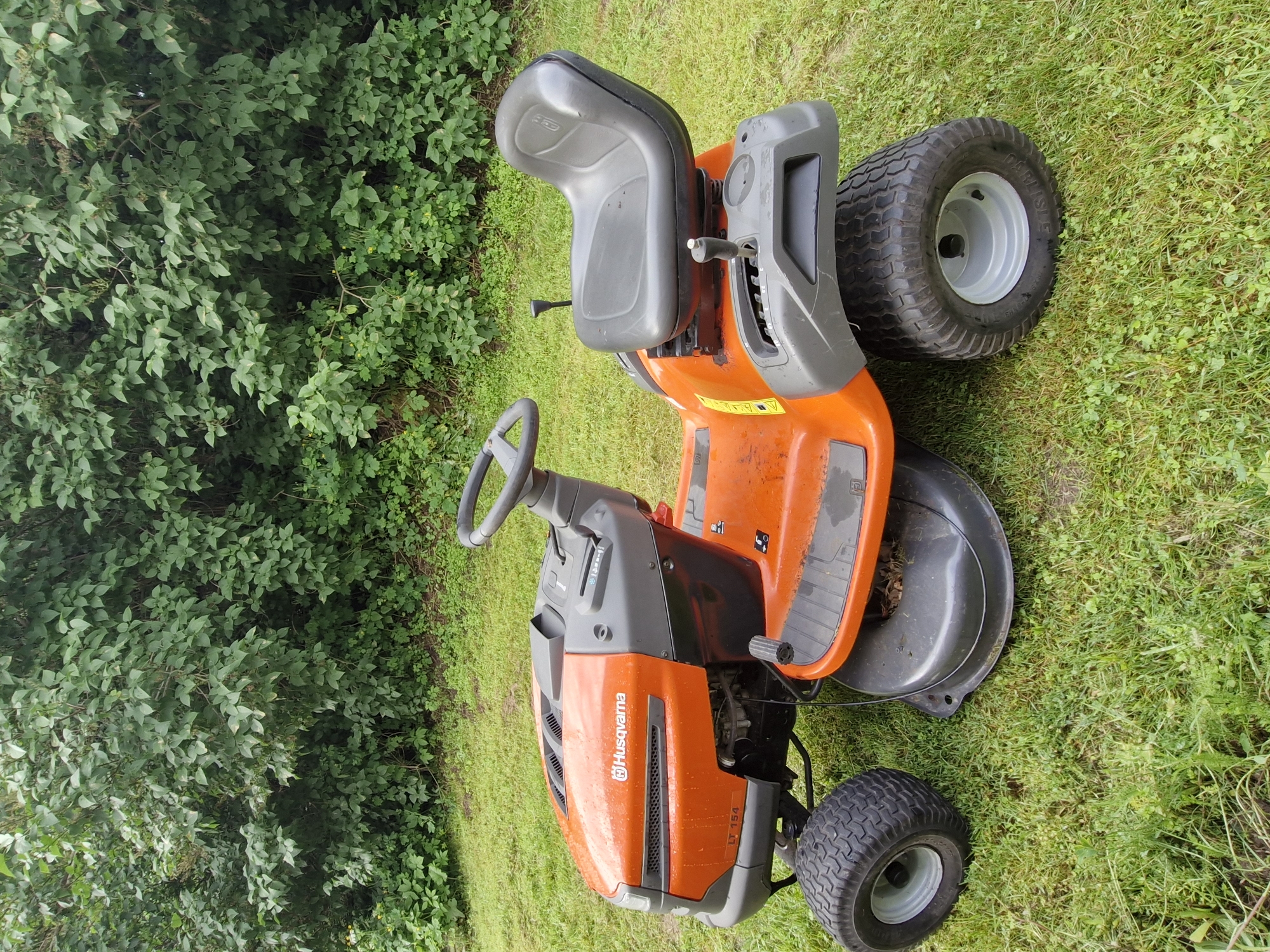 An orange and gray Husqvarna riding lawn mower, model LT 154, is parked on a grassy lawn in front of a dense green hedge. The mower is facing the right side of the frame. It has a black seat, a steering wheel, and large rear wheels with tread. A lever is visible near the seat. The grass is green and somewhat uneven. The background consists of lush, leafy green bushes and trees. The lighting suggests it is daytime, possibly overcast. There are no people visible in the image.