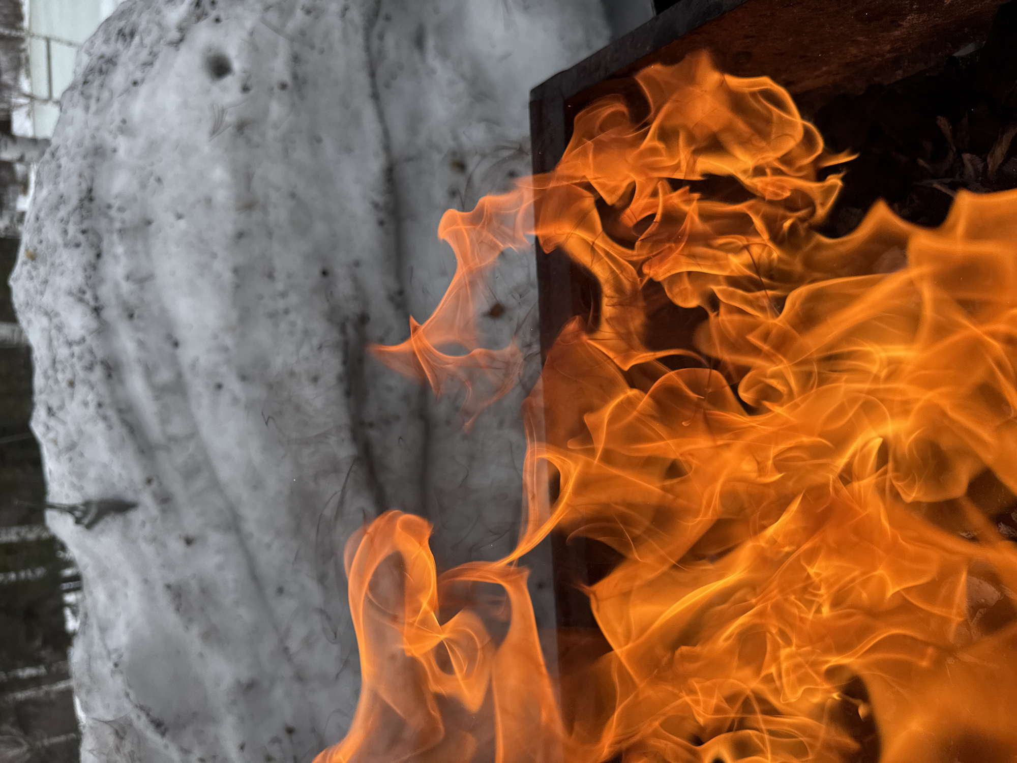 A close-up shot depicts a stark contrast between fire and snow. Intense orange flames fill the right side of the frame, originating from a dark, likely metal, container or structure. The left side presents a mass of snow, appearing dirty and partially melted. The photograph does not provide enough information to confirm the image's location in Votkinsk, Russia, or any other specific place. There are no people visible in the scene. The key elements are fire and snow in close proximity, suggesting a possible interaction or juxtaposition of the two contrasting elements.