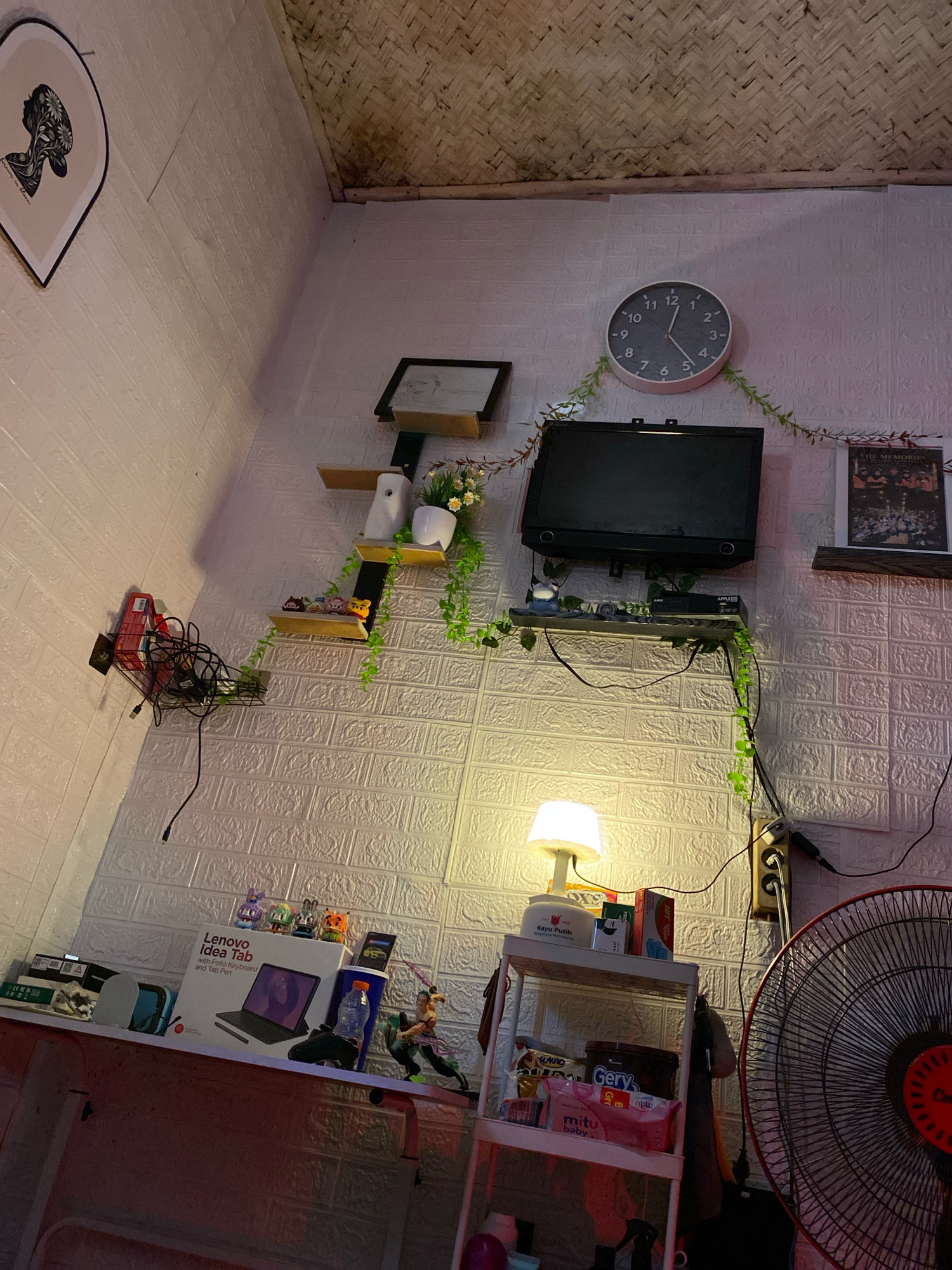 The image shows a dimly lit interior corner of a room, likely a bedroom or a personal study, in Hegarmukti, Indonesia. The walls are covered with white textured wallpaper, and the ceiling has a woven bamboo texture. On the wall, a round analog clock with black numbers and hands is prominently displayed, indicating approximately 11:53. Below the clock, a small black television is mounted, with a digital set-top box beneath it. Green artificial vines are draped around the television and extending down the wall, intertwined with wiring.
To the left, a framed artwork of a woman's silhouette with floral elements is hung on the wall. Further down the wall, a wire basket is attached, holding various electronic accessories, including cables and a red box.
Several small shelves are affixed to the wall. One shelf holds a white spray bottle, a potted plant with small white flowers, and a white pot. Another shelf below displays several small collectible figurines.
On a lower shelf, a Lenovo Idea Tab box is visible, along with other electronic accessories and a black game controller. A bottle of water and a toy figurine of a character with a sword are also present.
To the right, a white multi-tiered shelf unit is filled with various items, including packaged goods, a large tin, and a small lamp with a white shade emitting a warm glow. A power strip with multiple outlets is plugged into the wall, with several cords connected. In the foreground on the right, a large black oscillating fan is partially visible. The overall impression is of a cluttered but personalized living space.