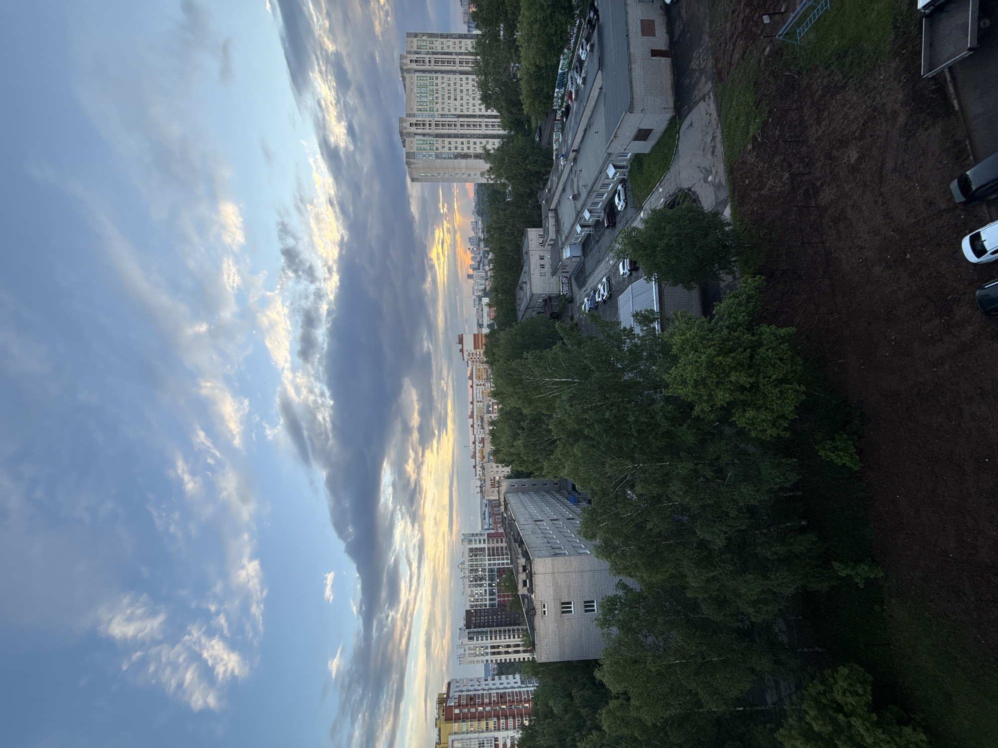 This image provides an elevated, wide-angle view of an urban landscape in Izhevsk, Russia, captured during either sunrise or sunset. The sky dominates the upper portion of the frame, showcasing a blend of clear blue and diverse cloud formations, from delicate white cirrus to broader, darker gray clouds. A prominent golden and orange light illuminates the horizon, where the sky meets a distant line of city buildings, signifying the early morning or late evening.
The scene features a mix of architectural styles, with tall, multi-story residential buildings, characteristic of post-Soviet design, standing prominently in the mid-ground and background. Lower-rise commercial or utility buildings are visible closer to the foreground, accompanied by several parked vehicles in adjacent paved lots. Extensive green spaces are interspersed throughout, with numerous mature trees, forming dense canopies particularly in the foreground and mid-ground, adding to the verdant feel of the area.
In the lower foreground, a large expanse of dark, disturbed earth suggests a cleared plot of land, possibly a construction site or undeveloped area, with patches of grass and a paved path running alongside it. A small blue structure, potentially scaffolding or a piece of playground equipment, is visible within this dirt area. Cars are parked on paved surfaces and what appears to be a dirt track. No people are discernible, and there is no visible text. The overall atmosphere is calm and peaceful, bathed in the soft, warm light of the golden hour.