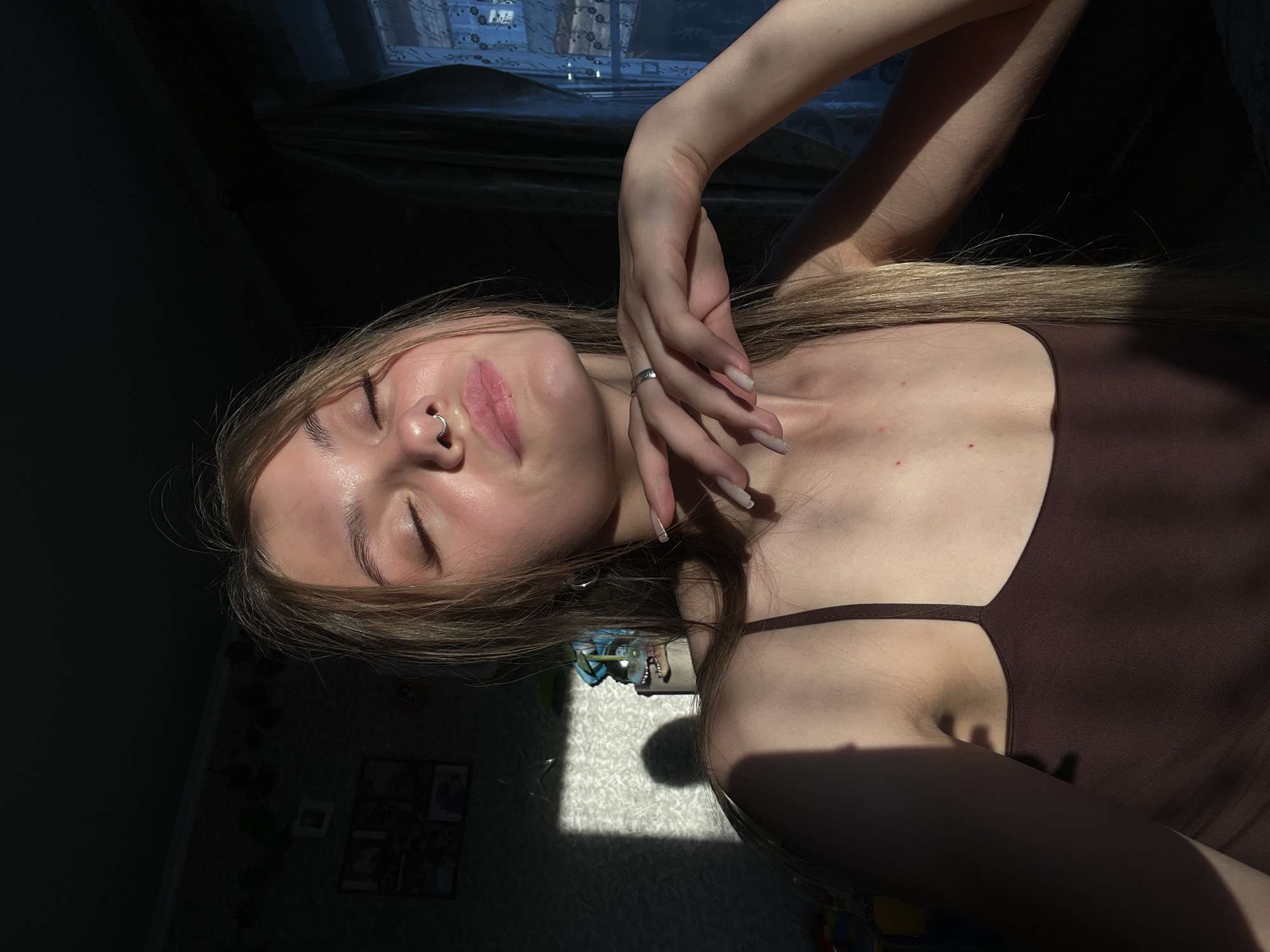 A young woman with blonde hair is pictured from the chest up, with her eyes closed and her head tilted back slightly. Sunlight is casting a strong shadow across the left side of her face and shoulder, highlighting her features. She is wearing a brown spaghetti strap top and has a septum ring and a ring on her left hand. Her right arm is bent, and her hand is resting on her chin. In the background, a dark wall with framed pictures is visible, as well as a window with blue curtains. The environment appears to be an indoor setting, likely a home, within the city of Fokino, Russia.