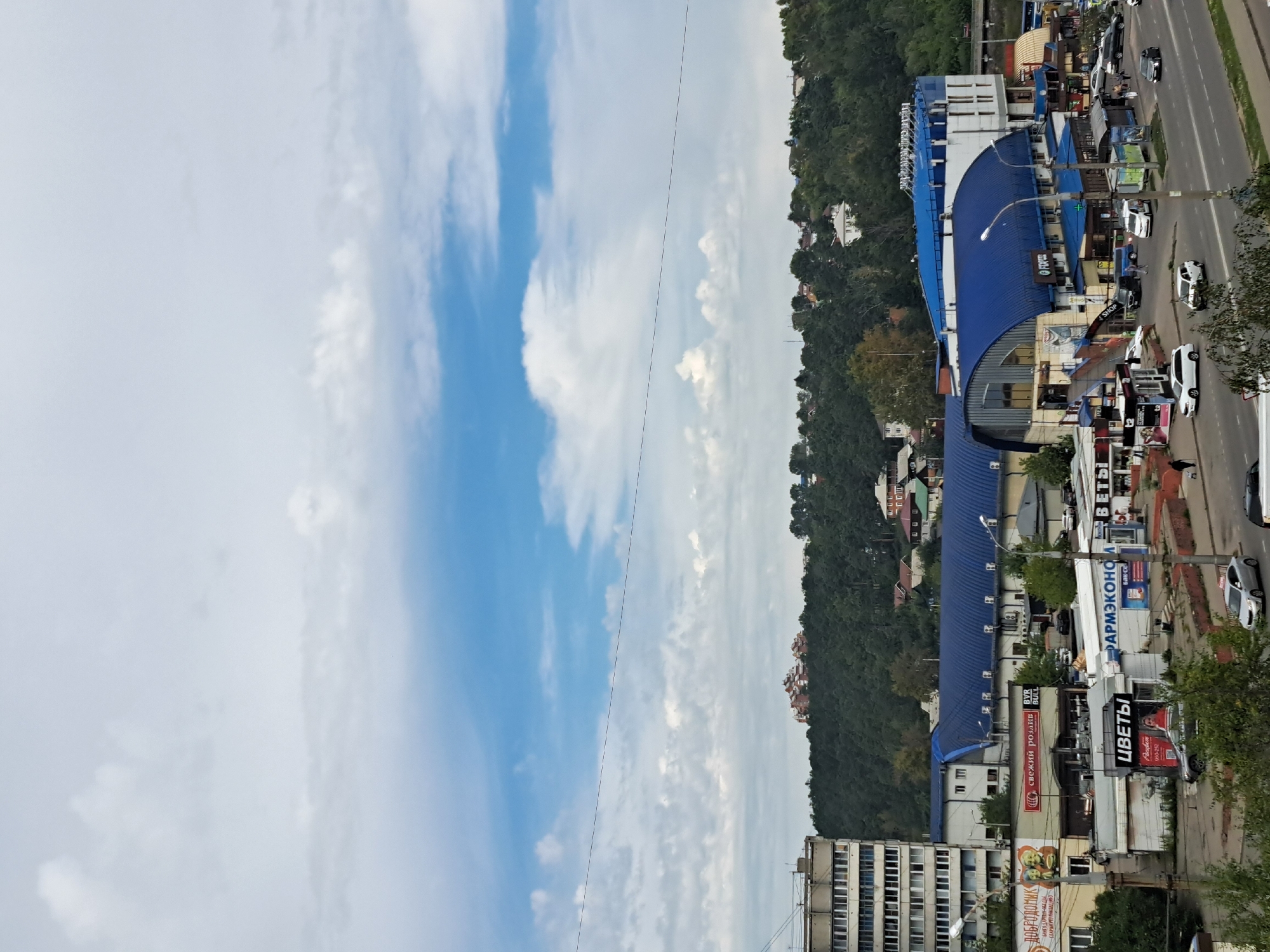 The image depicts an urban landscape with a commercial area in the foreground and a densely wooded hill in the background. The commercial area features a large building with a distinctive blue, curved roof, likely a shopping center or market. Several smaller storefronts are situated in front of this main building, some with prominent signage including "ЦВЕТЫ" (Flowers) and "ФАРМЭКОНОМ" (PharmEconomy). Cars are visible on the road and parked in the vicinity. The background is dominated by a steep, tree-covered hillside with scattered houses and buildings visible amongst the foliage. The sky is partly cloudy with a mix of blue sky and white clouds.
This image is a view of the Kachevskiy Market in Irkutsk, Russia. The market is situated at the foot of the Kachevskiy Hill, a prominent natural landmark in the city. The tall building on the left is likely an apartment block, a common sight in Soviet-era urban planning, which is characteristic of Irkutsk's architecture.