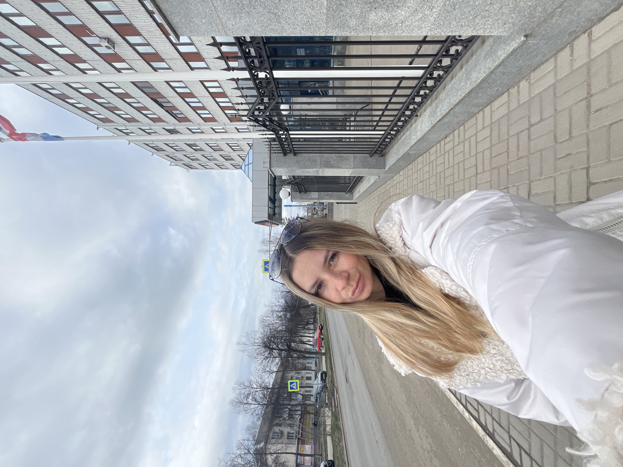 The image shows a woman in a white jacket standing in front of the administration building of the Chaikovsky urban district in Chaikovsky, Russia. The building has a multi-story facade with a repeating pattern of windows and a flag of Russia on a pole extending from the front. The sidewalk is made of light-colored paving stones and has a black metal railing. In the background is a street with trees and buildings, including a pedestrian crossing. The sky is overcast.