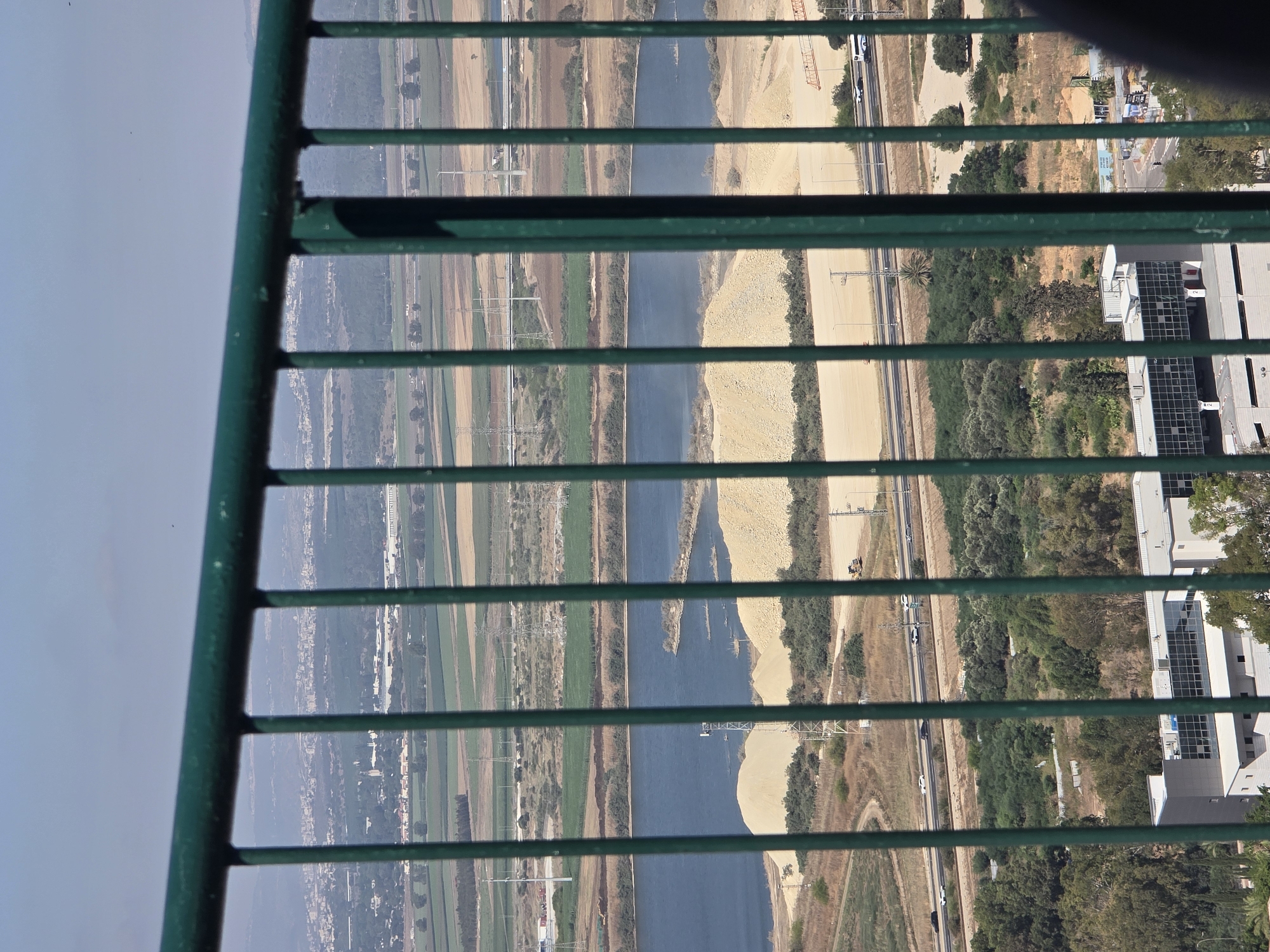 The image is a tilted, vertical shot looking down through a green metal railing. The scene is a wide landscape of a river, agricultural fields, a highway, and some buildings, seen from an elevated perspective. The river is a prominent blue feature, with sandy banks and vegetation on either side. Beyond the river, there are fields with varying shades of green and brown, suggesting different crops or stages of growth. A highway with cars is visible, running parallel to the river. In the background, there are some modern buildings with glass facades, one of which appears to be an industrial or commercial complex. There are also trees and some power lines or towers in the landscape. The time of day appears to be daytime, with clear skies, indicated by the bright sunlight and shadows cast by the railing. The overall impression is of an arid or semi-arid region with evidence of human activity and infrastructure interspersed with natural and agricultural elements. The location is identified as Nesher, Israel.