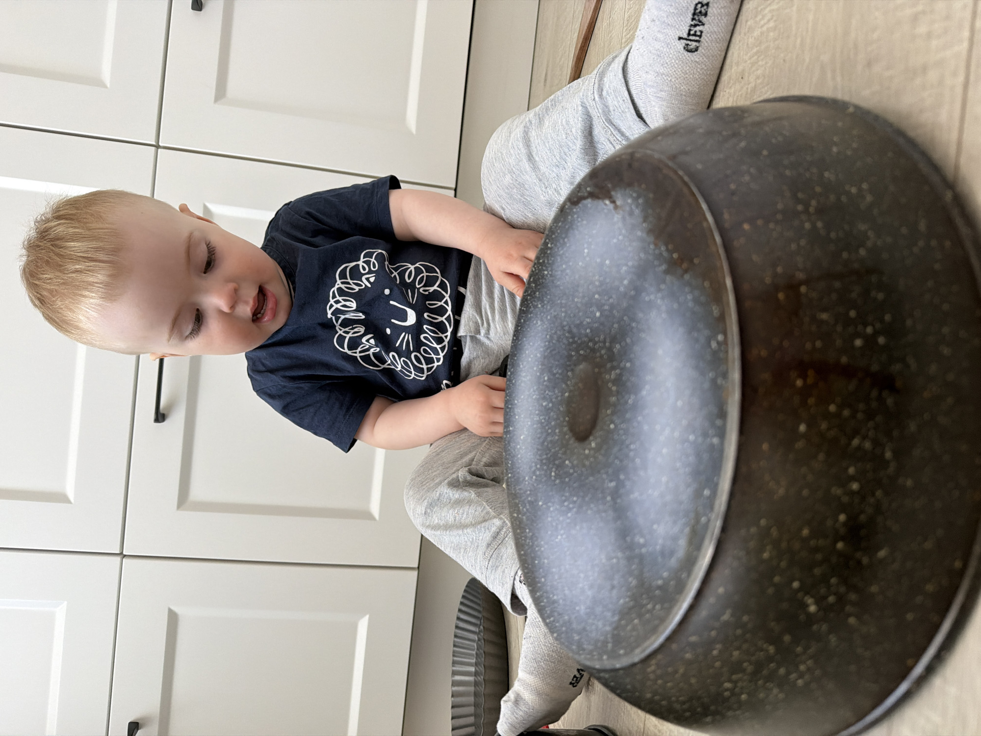 A toddler sits on a light wooden floor next to white kitchen cabinets. The child is wearing a dark blue t-shirt with a white graphic, gray sweatpants, and gray socks with black lettering. A large black frying pan is prominently positioned in the foreground, partially obscuring the child. A fluted metal tart pan is also visible on the floor. The location is inferred to be a domestic kitchen.