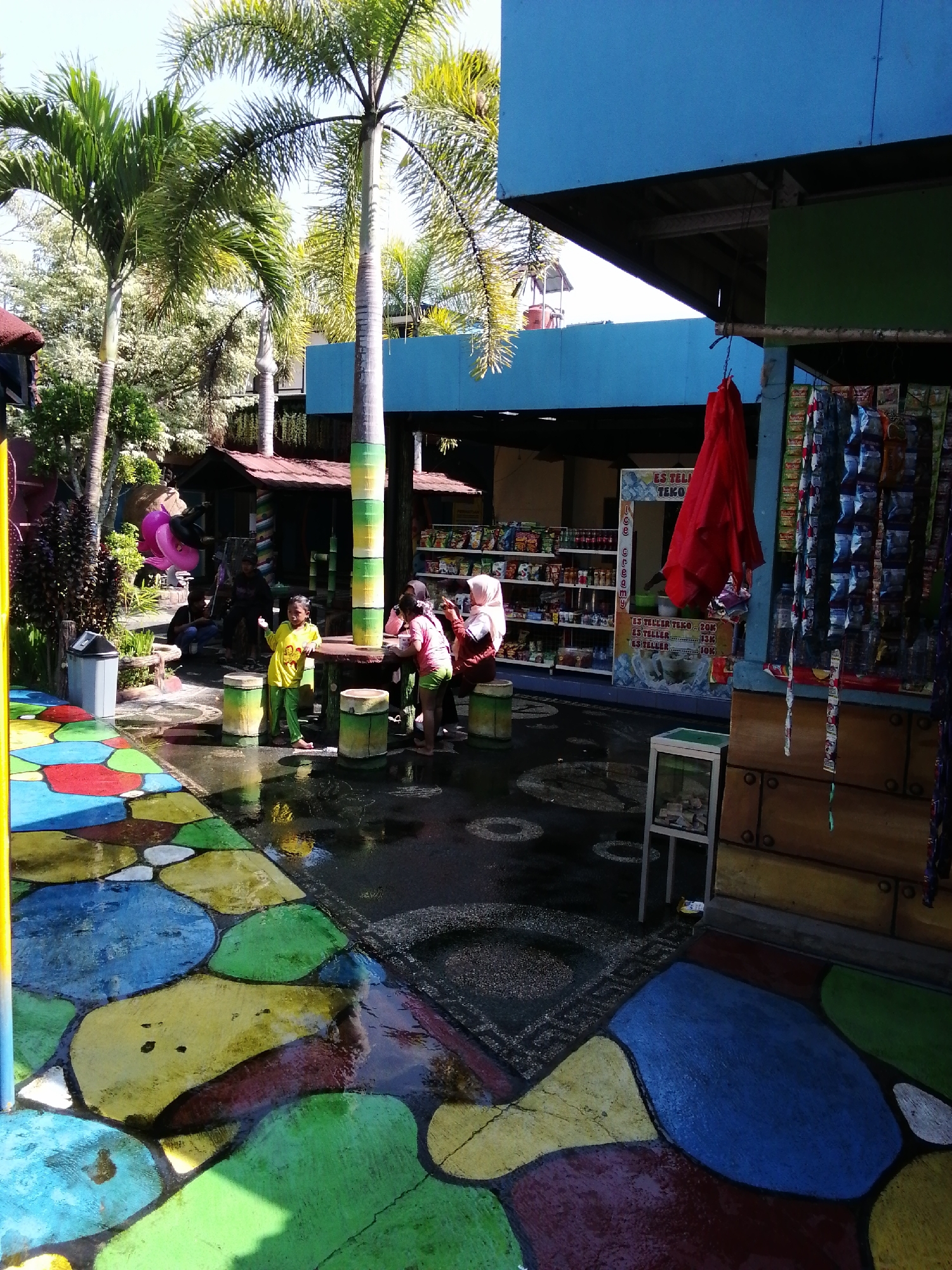 This image captures an outdoor scene at a water park in Tegalwangi, Indonesia, on a sunny day. In the foreground, a vibrantly colored pathway features large, irregular shapes in blue, green, yellow, and red, wet from recent activity. To the left, a gray trash bin sits next to a palm tree.
Further back, a group of people are gathered around a wooden table. A young girl in a yellow shirt and green pants stands near the table, appearing to interact with others. Other individuals, including women in hijabs and children, are seated or standing around the table. A prominent, tall palm tree with a green and yellow banded trunk stands near the gathering.
Behind the table area, a small building with a reddish-brown roof is visible, appearing to be a shop or food stall. Shelves stocked with various products, possibly snacks and drinks, are displayed outside this building, along with a red fabric hanging from a pole. A blue structure with a green panel extends into the scene from the right, and a small glass display cabinet with items inside is positioned on the ground. The overall environment suggests a recreational space, likely with water features, given the wet ground.