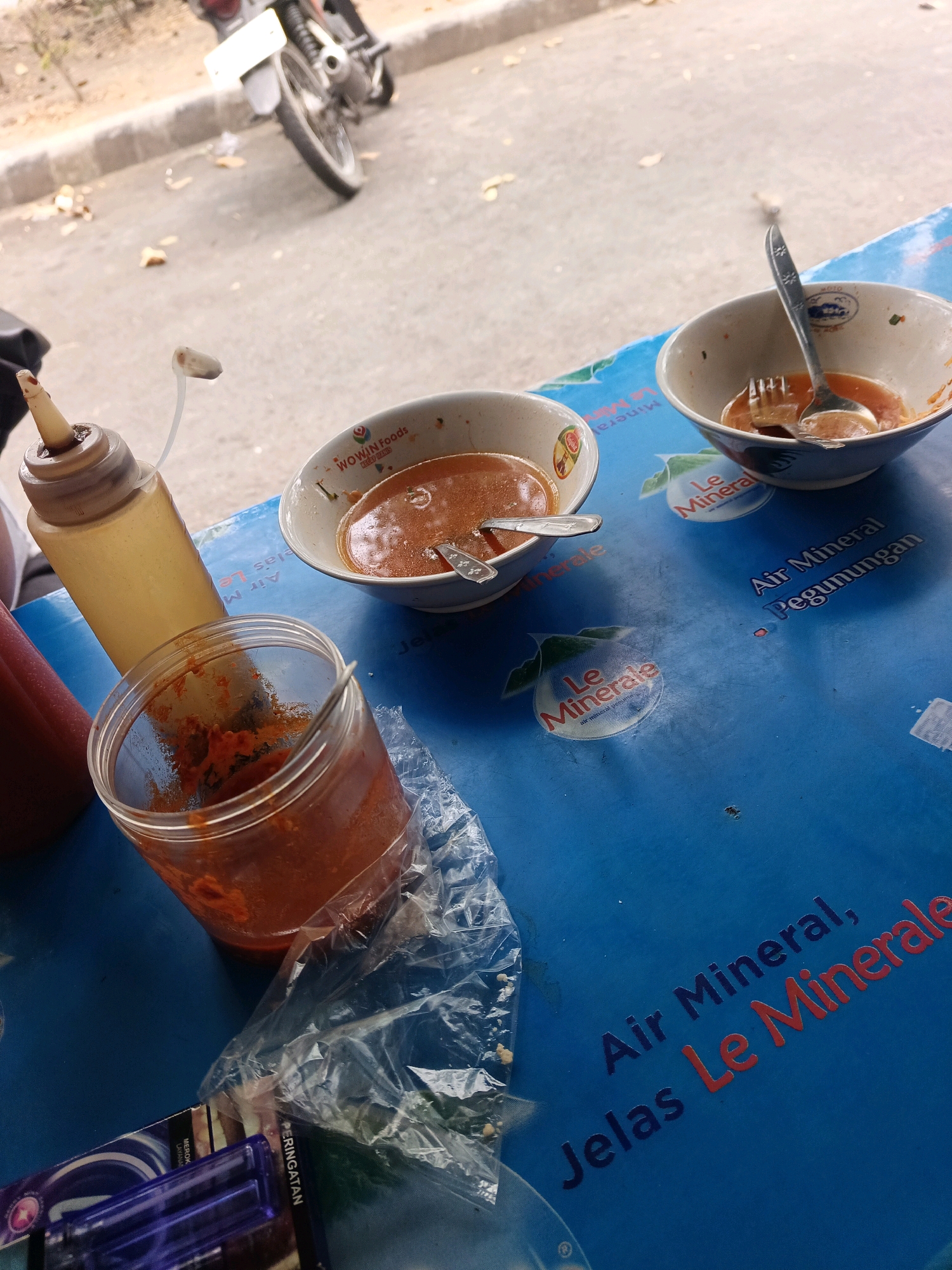A high-angle shot captures a blue plastic table in Kediri City, Indonesia, laden with food remnants and condiments. Two bowls containing a reddish sauce, presumably a savory dish, sit on the table, each accompanied by a spoon or fork. One bowl is partially visible in the foreground, while the other is further back, with cutlery resting inside. Adjacent to the bowls is a clear plastic jar filled with a similar reddish paste, likely chili sauce or sambal, also with a spoon. A yellow squeeze bottle with a white dispenser cap stands to the left of the jar, its contents unclear but possibly a sauce or condiment. To the far left, a red bottle, likely ketchup, is also present. In the background, a motorcycle is parked on a paved surface, its rear wheel and license plate visible. The table surface is adorned with text and logos, including "Le Mineral" and "Air Mineral Pegunungan," indicating the presence of bottled water branding. A crumpled plastic bag lies near the jar. The scene suggests a casual meal setting, possibly at an outdoor food stall or eatery. The overall impression is one of a meal that has recently concluded or is in progress, with the remnants indicating consumption.