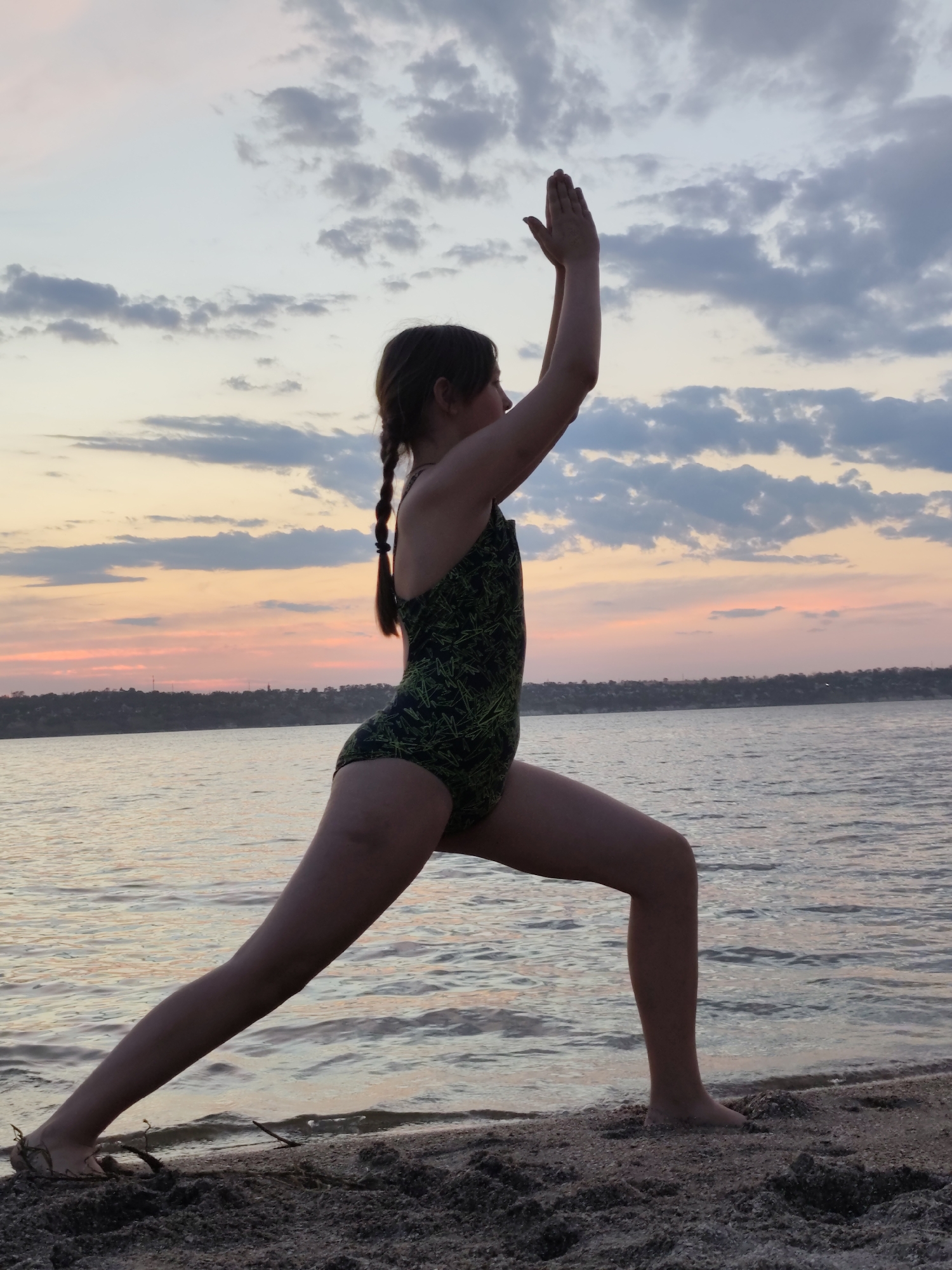 The image shows a young person practicing yoga on a sandy shore at dusk. The person is wearing a black swimsuit with a green leaf pattern and is in a Warrior II pose, with their arms extended and palms facing each other. Their left leg is bent, and their right leg is extended behind them. The person's hair is in two braids.
The setting is a beach with calm water, likely a river or sea, as indicated by the shoreline and the distant tree line on the opposite bank. The sky is filled with soft, colorful clouds, transitioning from pale blue to shades of pink and orange as the sun sets. The water reflects the colors of the sky, creating a peaceful and serene atmosphere. The overall mood is one of tranquility and mindfulness, enhanced by the natural beauty of the sunset and the calm setting. The location is Mykolaiv, Ukraine.