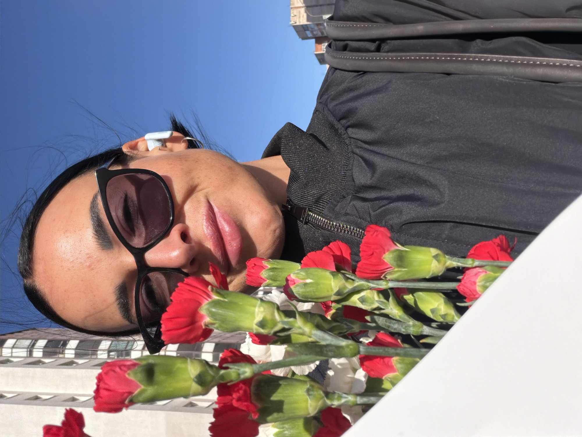 A medium shot shows a woman holding a bunch of red carnations, outdoors. The image is oriented at an angle, with the top of the frame showing a bright blue sky and the tops of buildings. The woman has dark hair pulled back, and is wearing sunglasses and what appears to be a wireless earbud. She is wearing a black jacket or coat. The carnations are predominantly red with green stems. The background behind the flowers appears to be white. The location appears to be an urban environment in Saint Petersburg, Russia, based on the architectural style of the buildings in the background.