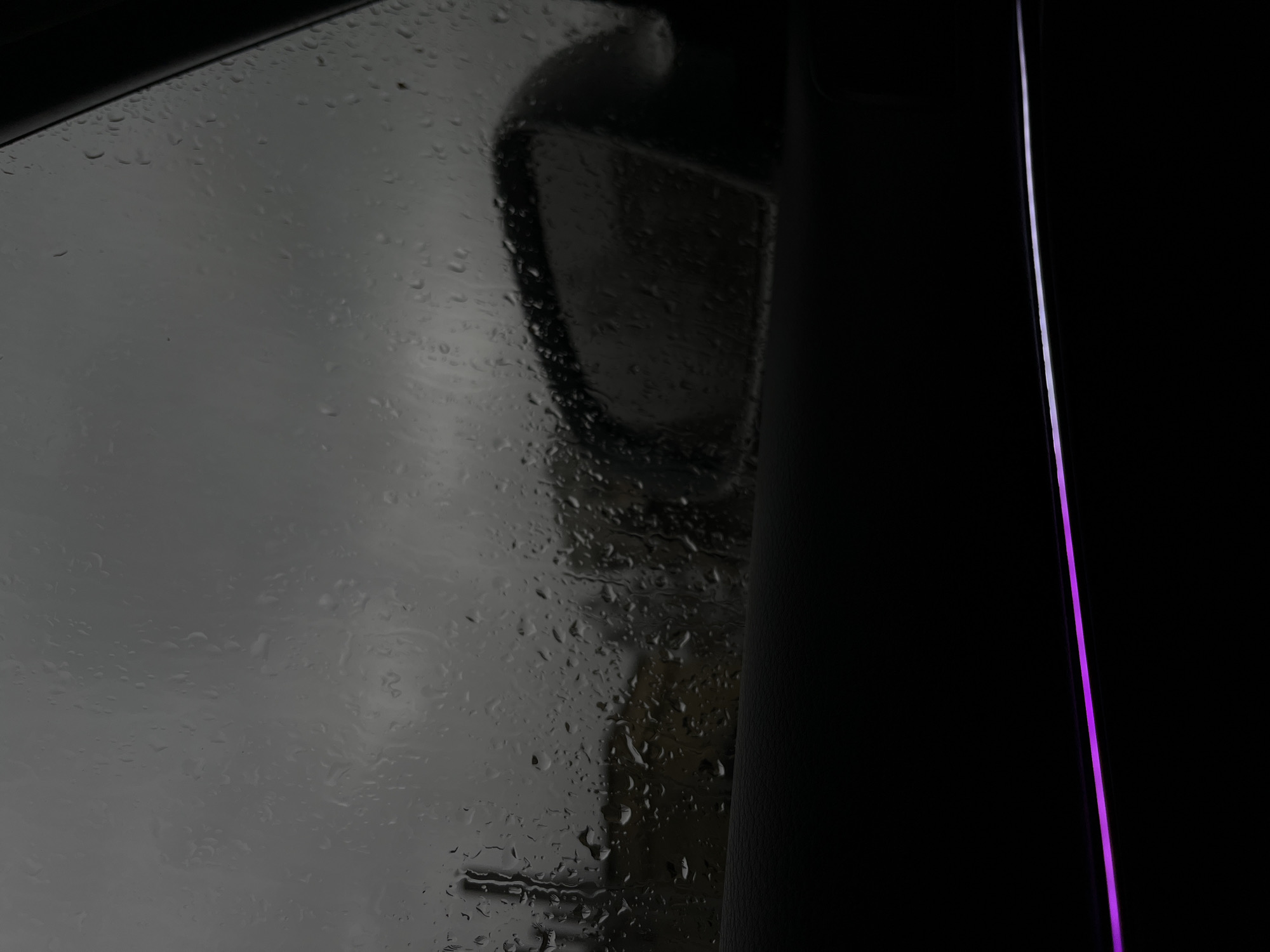 A close-up photograph shows a car window covered in raindrops. The reflection of a car side mirror is visible through the wet glass. The window frame is black and a thin vertical line of light, seemingly magenta, is present in the right part of the frame. The outside scene is blurred by the rain, making it difficult to identify specific locations or objects in Vratsa, Bulgaria.