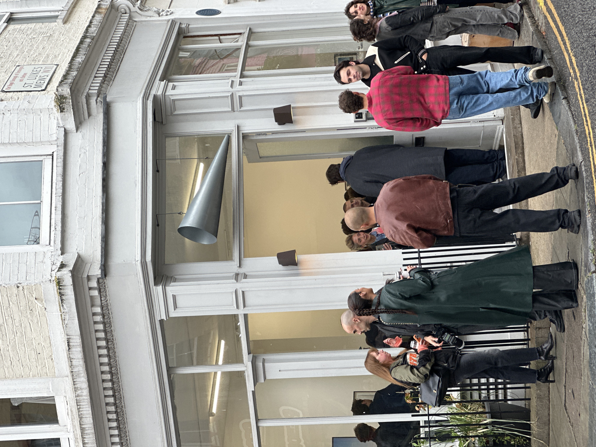 A group of people are gathered outside a building with a modern facade in London, United Kingdom. The building has large glass windows and a contemporary design. A street sign on the corner of the building reads "ST. LUKES MEWS, W.11". Several individuals are dressed in casual attire, and some appear to be holding drinks or cameras, suggesting a social gathering or event. The sidewalk and street in front of the building are visible, with yellow lines marking the road. The overall impression is of an urban street scene during daytime.