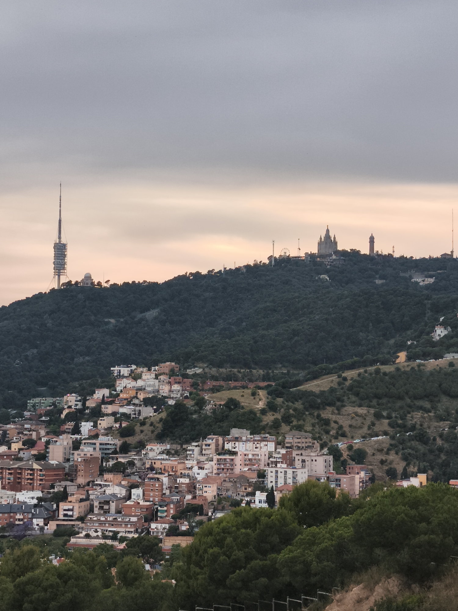 The image shows a panoramic view of Barcelona, Spain, at dusk. In the foreground, there is a cluster of residential buildings with pink and white facades, interspersed with trees. Behind these buildings, a large, densely forested hill rises prominently. Atop the hill, there is the Temple Expiatori del Sagrat Cor, a Roman Catholic church, and a television tower, Torre de Collserola. The cloudy sky suggests it might be early morning or late evening. There is no evidence of people in the image.
