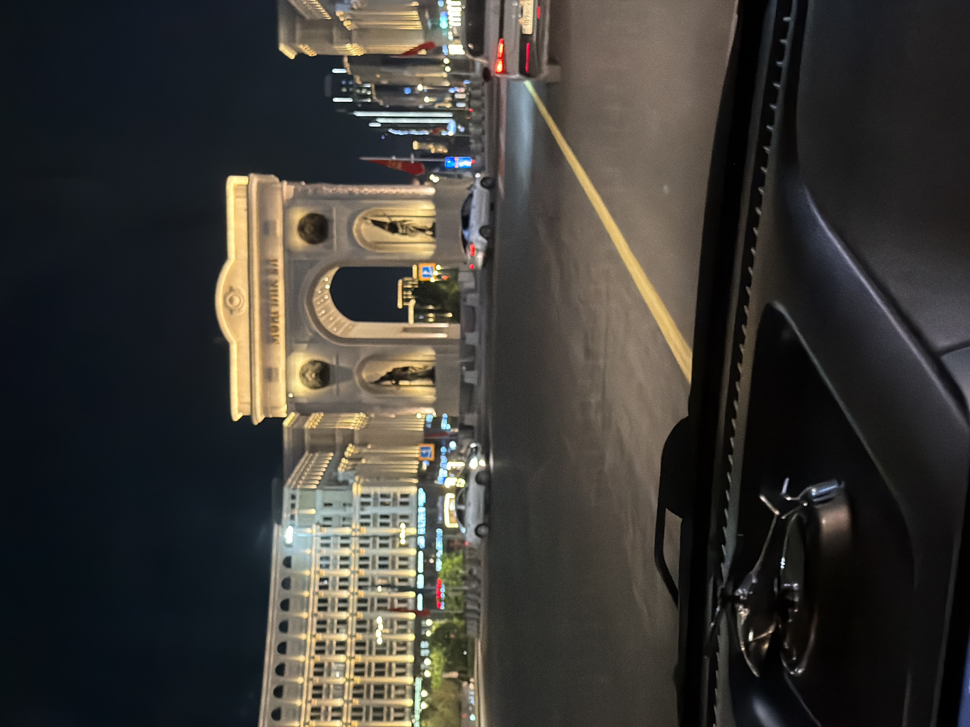 The image captures a nighttime view from a vehicle in Astana, Kazakhstan, looking towards a grand triumphal arch illuminated by warm lights. The arch features intricate architectural details, including statues and decorative elements, with Cyrillic text visible above the archway. Flanking the arch are modern buildings, also lit up, contributing to a city-like atmosphere. Several cars are visible on the road, indicating traffic. In the foreground, the dashboard of the vehicle is partially visible, featuring a metallic helicopter ornament. The overall scene suggests a well-lit, urban environment at night.