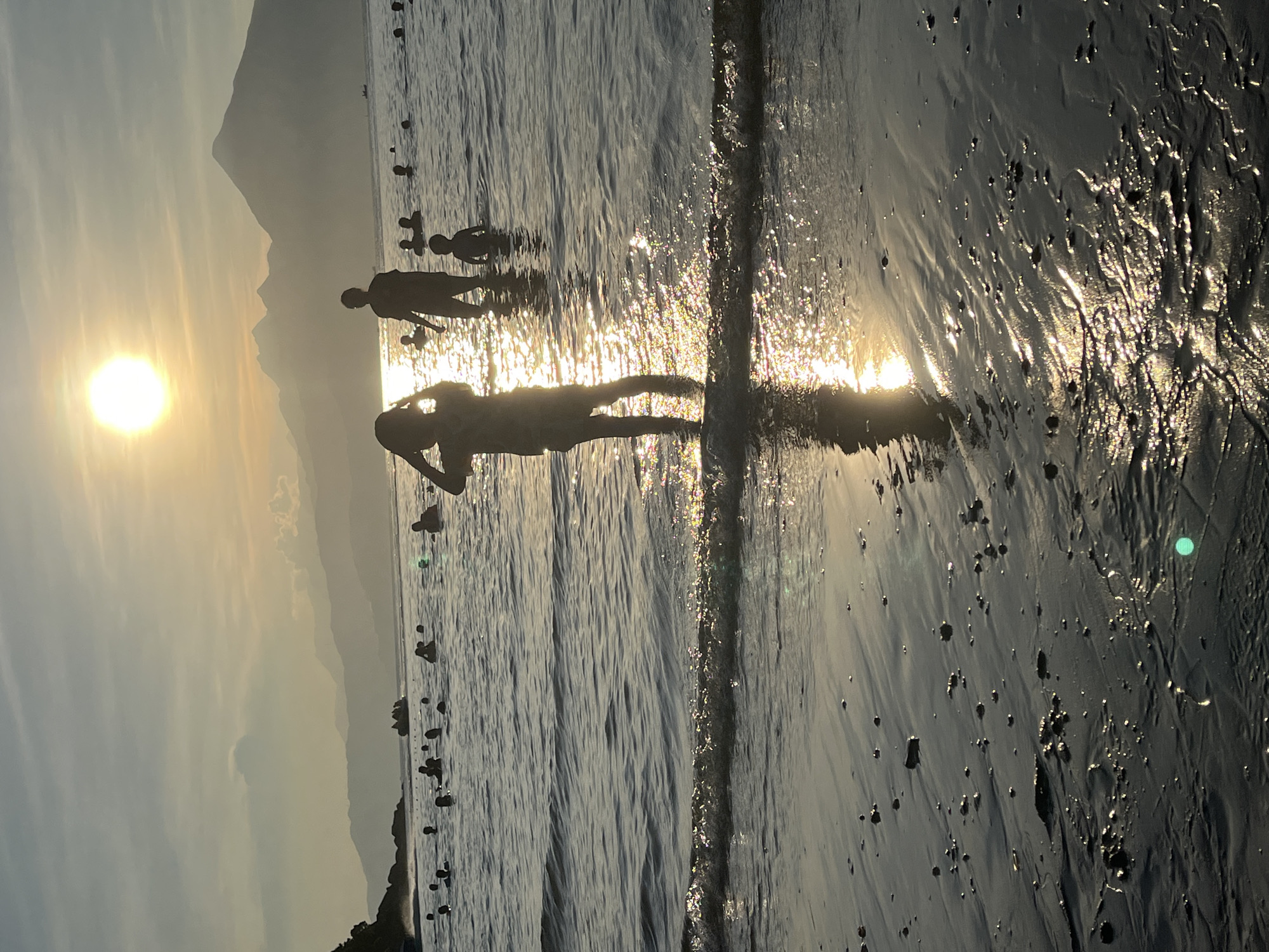 The image displays a tranquil beach scene at sunset or sunrise, dominated by strong backlighting from a low-lying sun. The bright, circular sun is positioned towards the left of the frame, illuminating the sky with a golden hue that fades into soft blues. Its intense light creates shimmering reflections on the surface of the shallow ocean water and wet sand, forming a sparkling path towards the viewer.
Several silhouettes of people are visible, engaging in leisure activities. In the foreground, a person stands in the shallow water or on the damp sand, with their reflection clearly visible. Further into the water, another person stands, and to their right, a small group of figures, likely children, appear to be wading or playing. Additional faint silhouettes are scattered across the water's surface, suggesting other beachgoers. Small, dark, rounded objects, possibly buoys or rocks, are interspersed throughout the water.
In the background, a range of hazy mountains stretches across the horizon, providing a dramatic backdrop to the coastal setting. The water appears calm with subtle waves breaking near the shore, contributing to the reflective quality of the surface. The overall atmosphere is peaceful and recreational, capturing the beauty of the golden hour at a scenic beach, consistent with locations like Đà Nẵng, Vietnam. No visible text is present in the image.