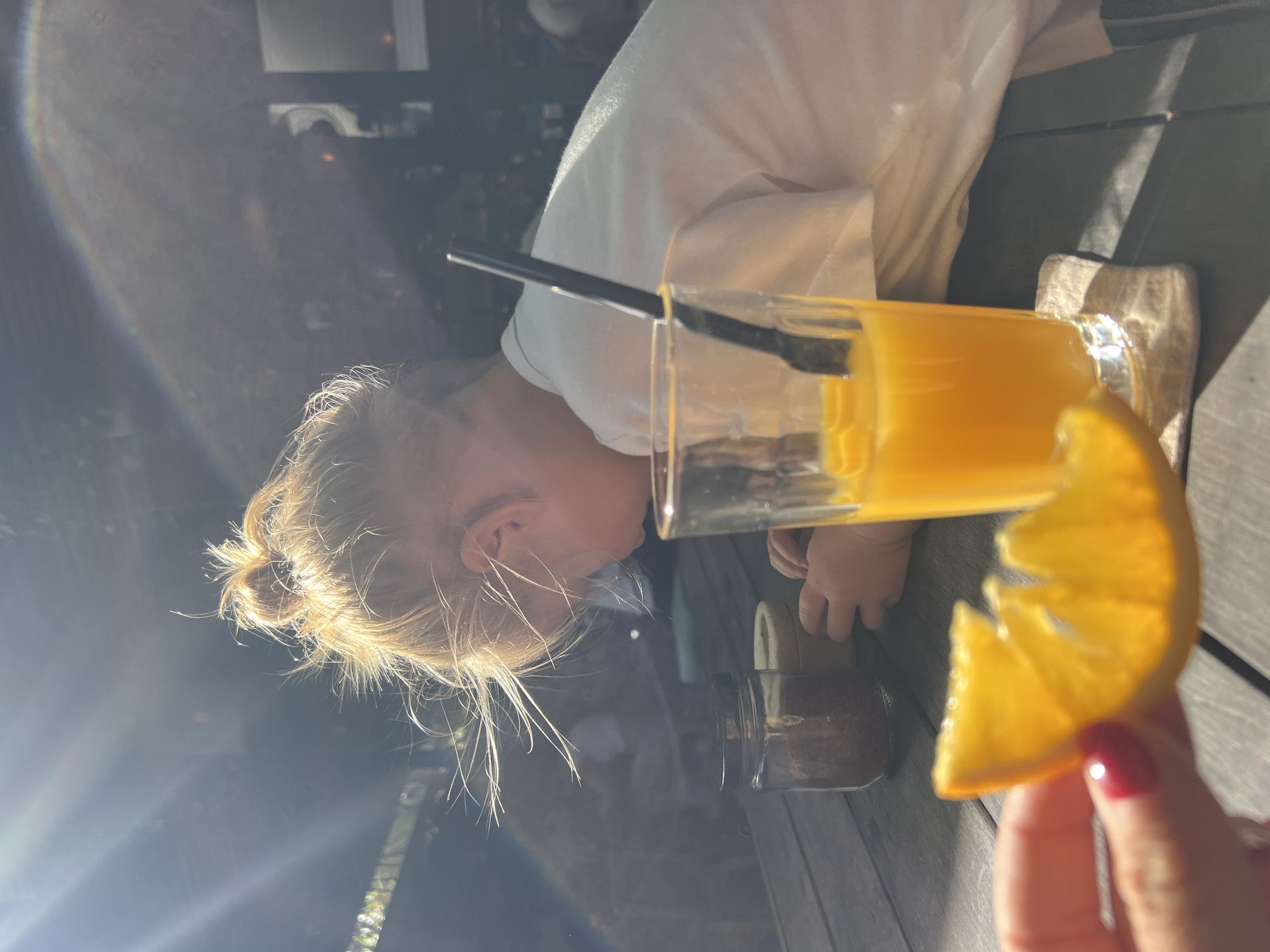 In North Kuta, Indonesia, a toddler with blonde hair pulled into a messy bun is seated at a wooden table, facing away from the camera and towards the right. The toddler is wearing a white t-shirt and appears to be reaching towards two small jars on the table. In the foreground, a hand with red nail polish holds a slice of orange. To the right of the toddler, a tall glass filled with orange juice and a black straw sits on a coaster. The scene is lit by strong, dappled sunlight, creating lens flare and casting shadows on the table. In the background, out-of-focus elements suggest an outdoor or semi-outdoor setting.