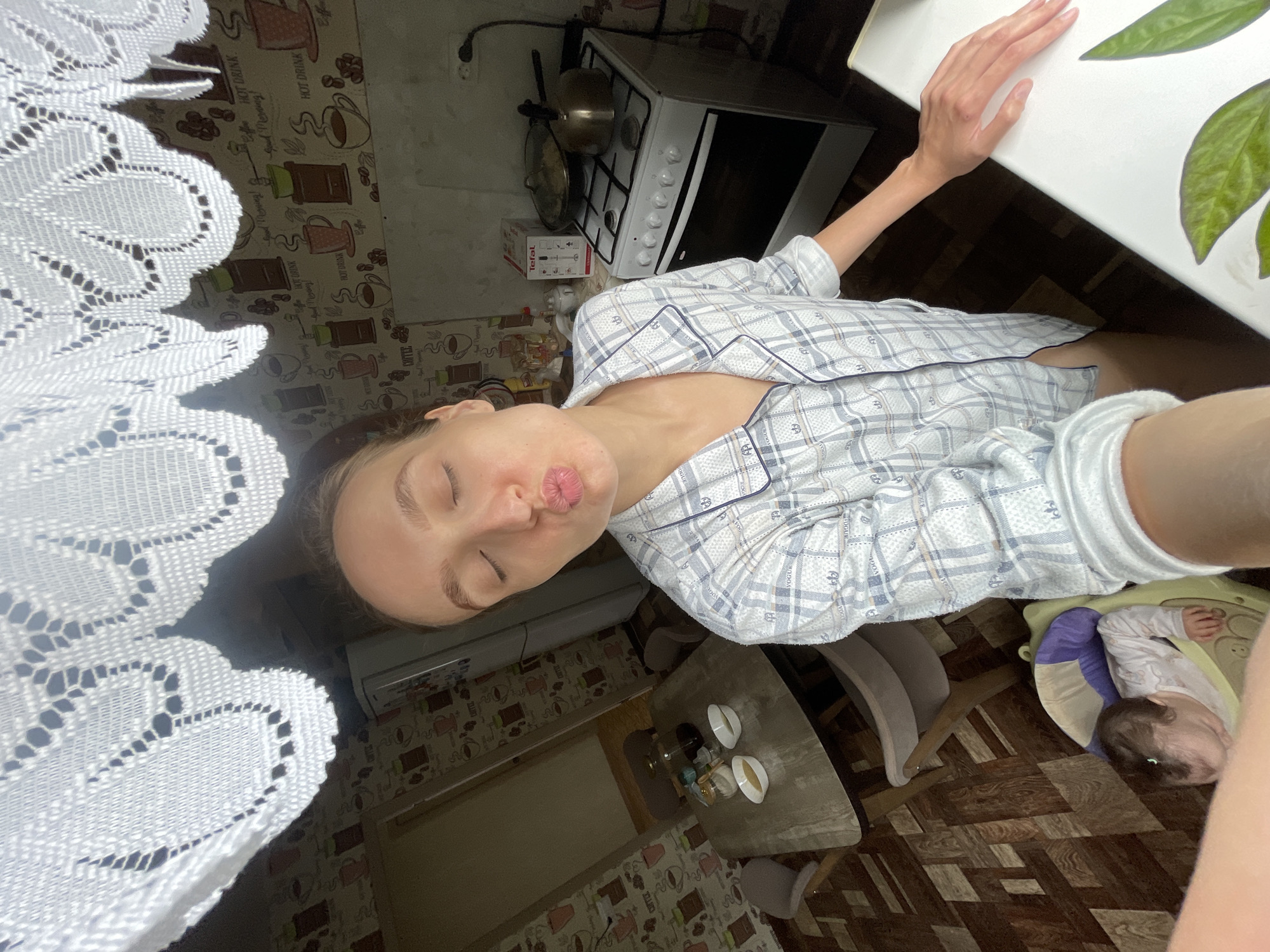 A selfie taken indoors by a young woman with her eyes closed and lips puckered in a kiss. She is wearing a blue and white plaid pajama top. In the background, a kitchen is visible with a stovetop, a kettle, a Tefal immersion blender box, and a table with bowls and jars. A baby is in a bouncy seat on the floor in the lower left corner of the image. The wallpaper has a coffee cup and coffee bean pattern. The location is the city of Sharkan, Russia.