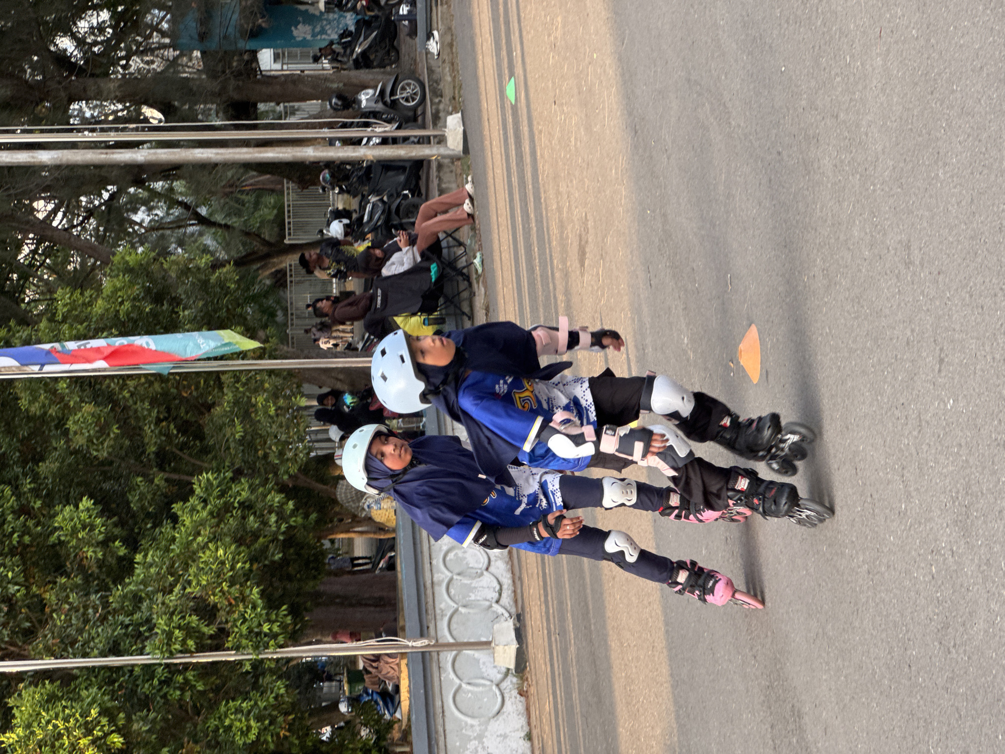 Two young girls, dressed in blue athletic wear and white helmets, are inline skating on an asphalt path in Banda Aceh, Indonesia. The girl in the foreground is wearing protective padding on her knees and elbows, as is the girl behind her. They are skating in the same direction, and the girl in the front appears to be looking towards the left of the frame. In the background, several people are seated on chairs, some appearing to be watching the skaters. Motorbikes are parked behind the seated individuals, and trees line the area. A flag with red, white, and blue colors is visible in the upper left of the image. The lighting suggests it is daytime, with shadows cast on the ground.