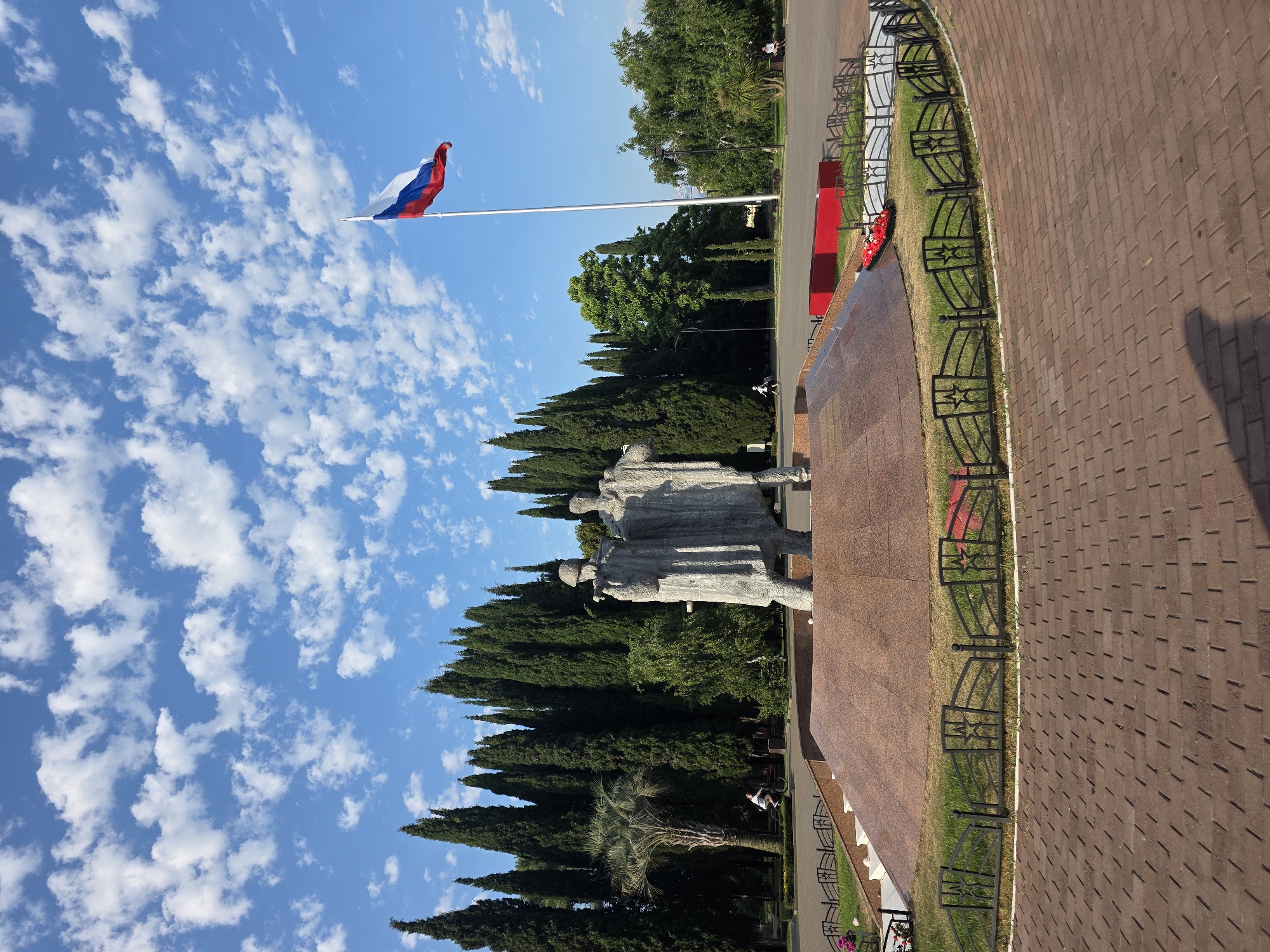 The image depicts a monument to two figures in Sochi, Russia, located in front of a row of tall cypress trees. The monument is made of stone and stands on a circular, multi-tiered base covered in red granite. A Russian flag flies from a tall flagpole to the right of the monument, against a backdrop of a bright blue sky with scattered white clouds. In the foreground, a circular brick pathway leads towards the monument. A black decorative fence with star motifs encloses the grass area around the monument. A person is visible cycling in the background on the left, and another person is sitting on a bench on the right. The overall scene suggests a public park or square.