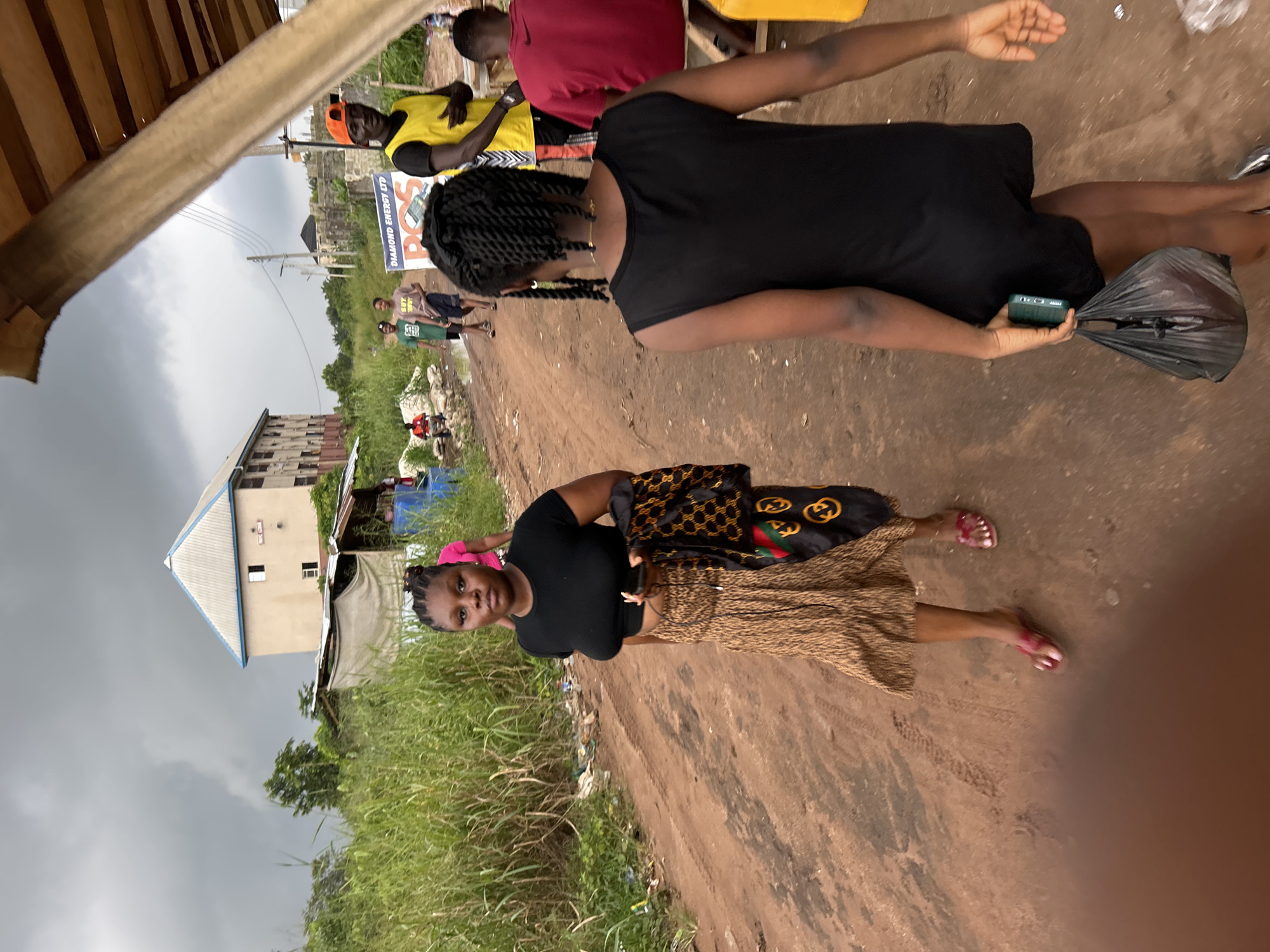 The image shows a group of people in what appears to be a rural or semi-urban setting in Ihiagwa, Nigeria. In the foreground, a young woman with braided hair is wearing a black top and a patterned skirt. She is holding a black plastic bag in her right hand and a smartphone in her left. Behind her, another young woman is visible from the shoulders up, wearing a black tank top. Further in the background, several other individuals are present. One man is wearing a bright yellow vest over a black shirt, and another person is wearing a red shirt. A building with multiple stories and a distinctive roof is visible in the background, along with lush green vegetation and a dirt road. The sky is overcast, suggesting a cloudy or possibly rainy day. A sign with the text "DIAMOND ENERGY LTD" is partially visible. The overall scene depicts everyday life in a Nigerian community, possibly a marketplace or a residential area.