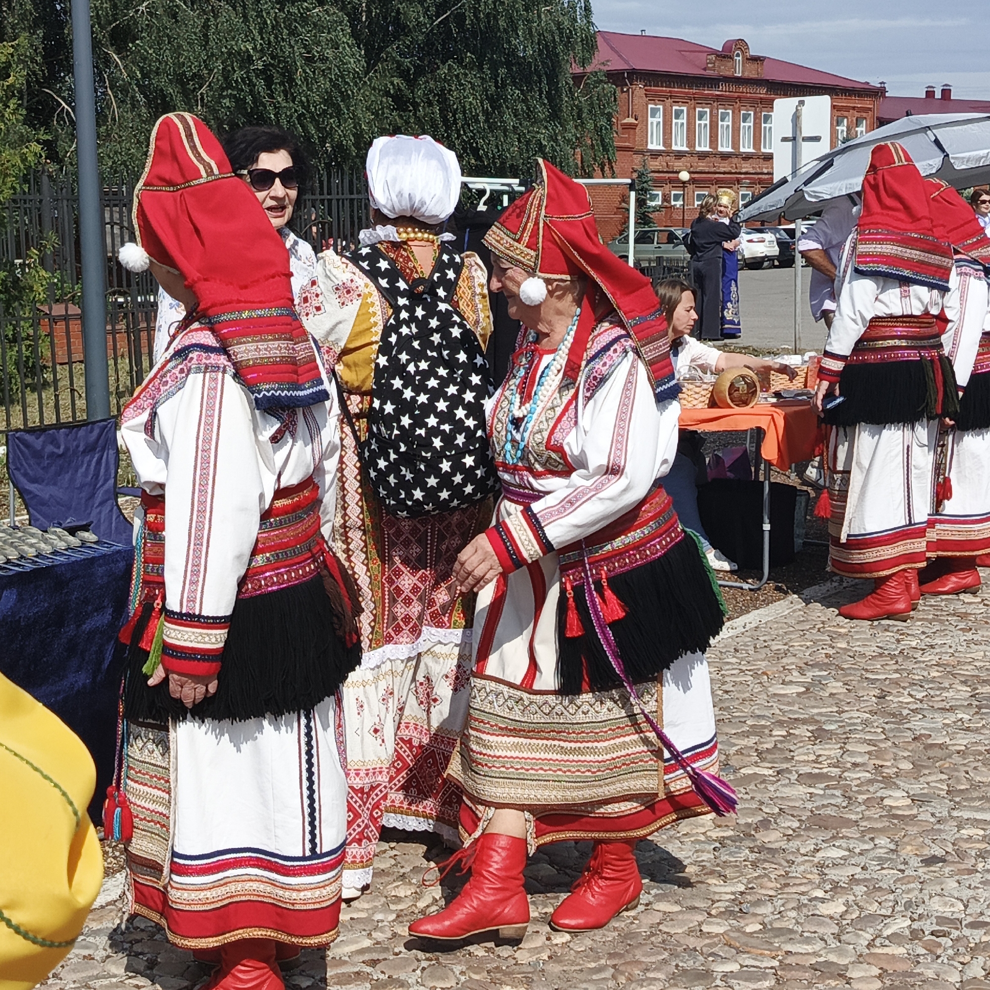 In the city of Nikolo-Beryozovka, Russia, on a cobblestone street, several people are gathered. The focal point is a group of women dressed in traditional folk costumes. These costumes are predominantly white with elaborate red and multicolored embroidered patterns on the sleeves, bodices, and skirts. They also feature black fringe accents and red boots. One woman in the foreground wears a red headdress adorned with a white pom-pom. Another woman to her left has a white headdress and wears a black garment with white stars. Behind them, other individuals are visible, some in similar attire, while others wear more contemporary clothing. In the background, a large, red brick building with white-trimmed windows stands under a clear blue sky. A black metal fence lines the left side of the scene, and a blue tarp covers a table with items displayed on it. A white umbrella provides shade for a section of the gathering. The overall atmosphere suggests a cultural event or festival.