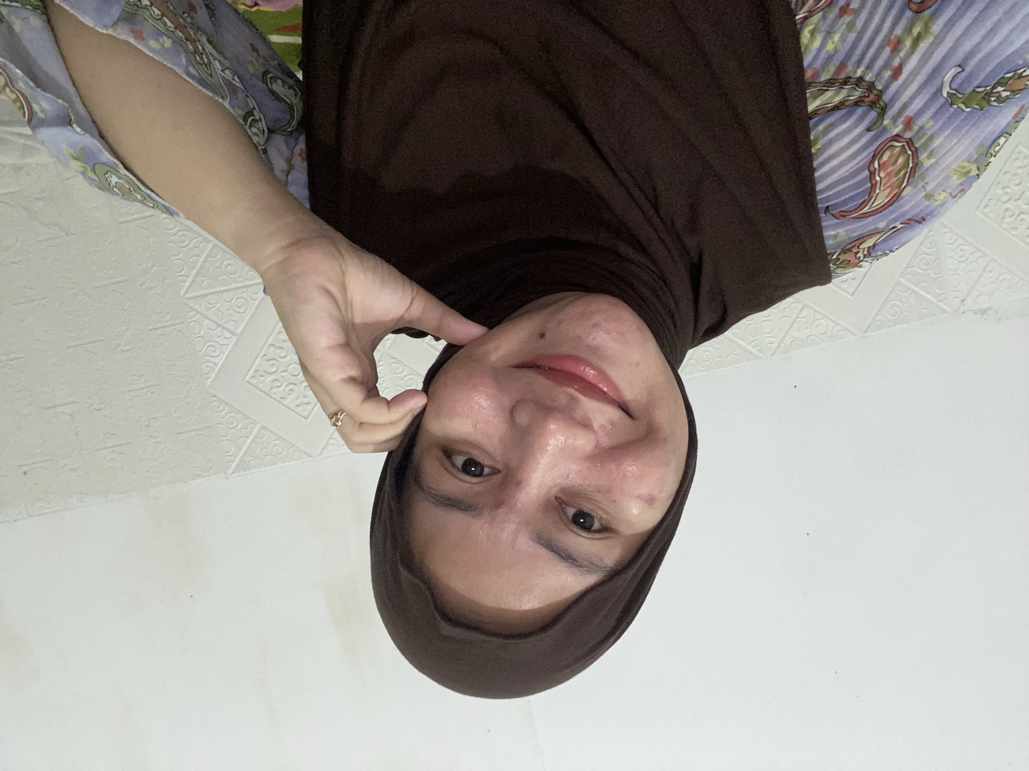 The media file is a vertically oriented, close-up selfie of a person wearing a dark brown hijab. The subject's face is turned towards the camera, and their right hand is touching their cheek, with a ring visible on their ring finger. The person has clear skin with some blemishes and is wearing lip gloss. Their eyes are dark and they are smiling subtly.
The background appears to be a white, textured wall, possibly with a patterned wallpaper. The lighting suggests an indoor setting, likely during the daytime given the brightness. The subject is also wearing a patterned garment on their left shoulder, featuring paisley designs in shades of blue, purple, and red.
The image is taken in Banjarbaru, Indonesia. There is no visible text in the media file. The overall impression is a casual, self-portrait, conveying a sense of pleasantness and engagement with the viewer.