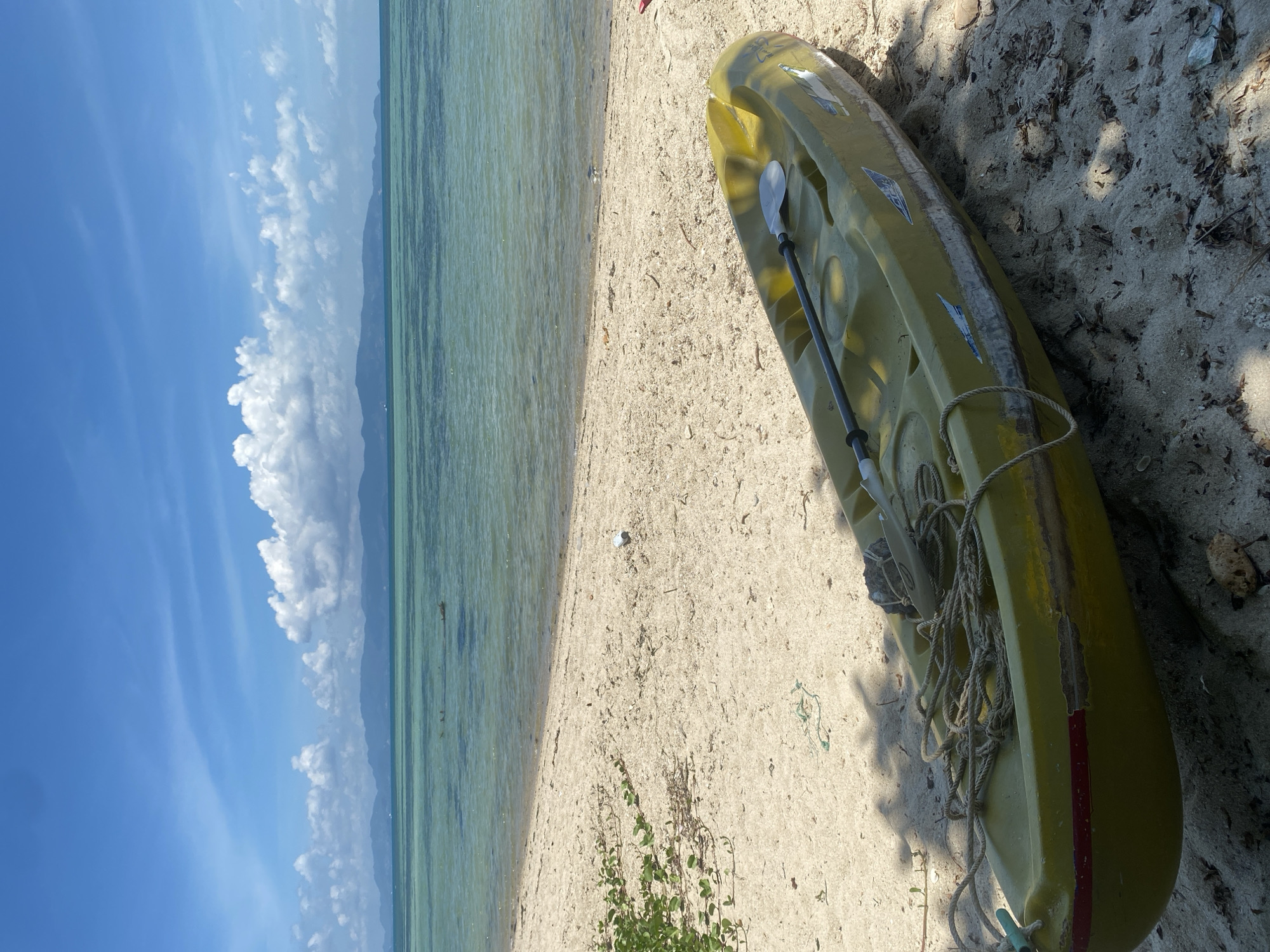 This image depicts a serene tropical beach scene in Moo 1, Thailand, under a clear, bright blue sky dotted with fluffy white clouds. The photograph is oriented vertically, showcasing a sandy shore, the clear turquoise sea, and distant land.
In the foreground, a lime green and yellow kayak rests on the light-colored sand, angled towards the water. A paddle is laid across the top of the kayak, and several coils of rope are gathered near its stern. The kayak shows signs of wear, with visible scratches and patches, and a red stripe near its tail. Shadows cast by the kayak and unseen foliage suggest bright sunshine, likely late morning or early afternoon. Small green plants and some scattered debris are visible on the sand near the water's edge and around the kayak.
The calm, shallow sea stretches across the midground, exhibiting clear, translucent waters with shades of green and blue. In the background, a low-lying range of mountains or distant land mass is visible across the horizon, partly obscured by the cloud formation. No people are present, contributing to the tranquil and undisturbed atmosphere of the beach. There is no visible text in the image.