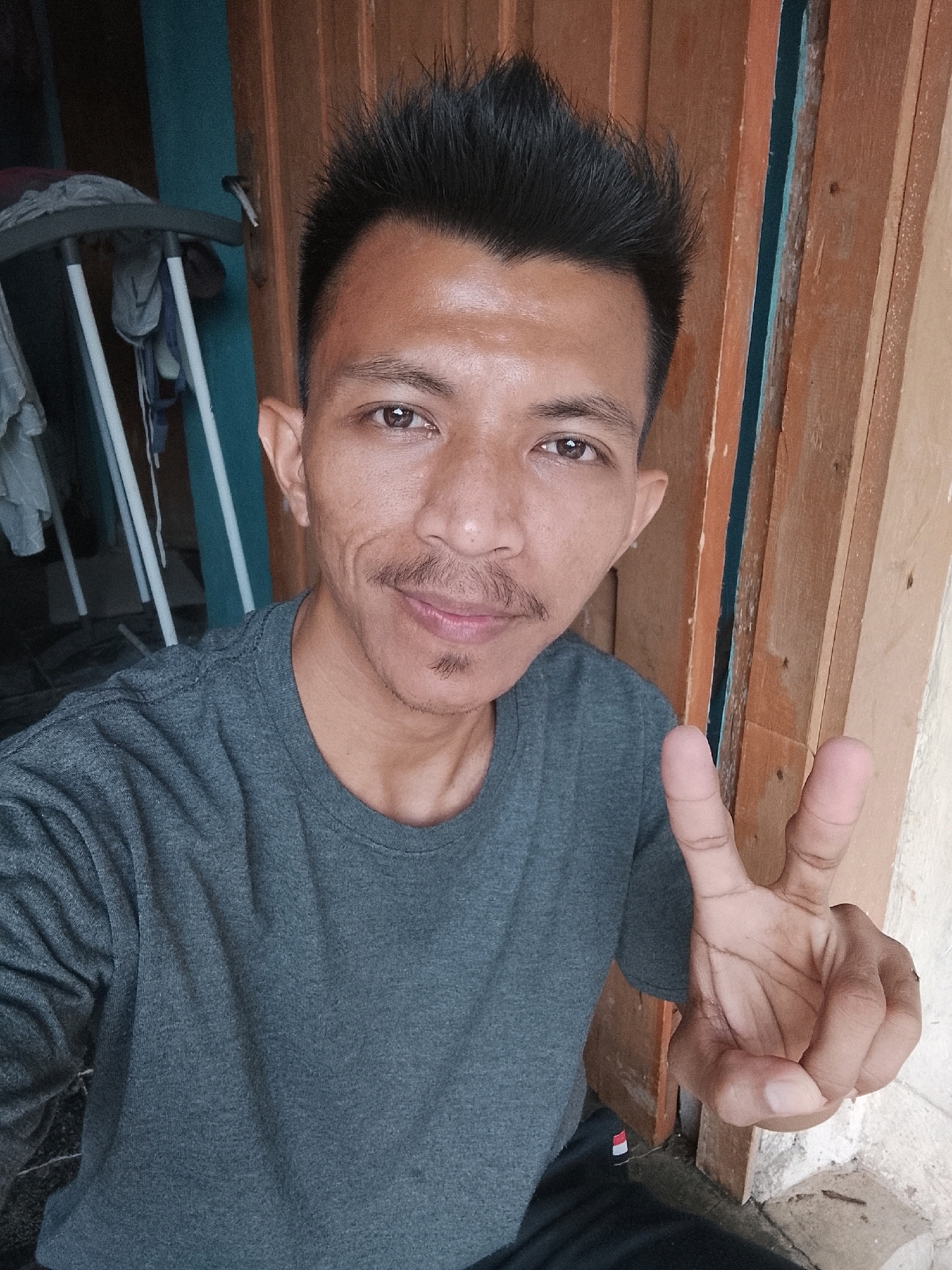 A medium shot captures a man in his late twenties or early thirties, with dark, spiked hair and light brown skin. He is wearing a dark grey t-shirt. He is holding up his right hand in a peace sign. The background consists of a wooden door frame to his right and a rack with clothes behind him to his left. The man is looking directly at the camera, with a slight smile. The location appears to be outdoors, possibly in a residential area, given the wooden door and the simple rack of clothes in the background. The overall impression is casual and informal. The image does not provide specific details to pinpoint its location within Kertahayu, Indonesia.