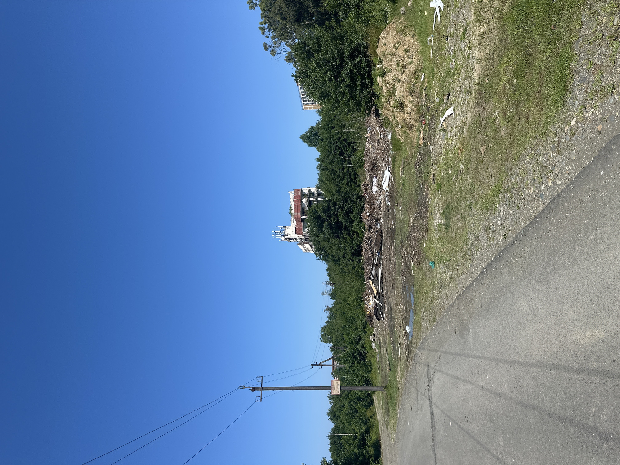 In Ureki, Georgia, an outdoor scene depicts a clear blue sky above a landscape with signs of abandonment and neglect. In the foreground, an asphalt road runs diagonally from the lower left to the mid-right of the frame. To the right of the road, a patch of overgrown grass and weeds is littered with debris, including pieces of white plastic, wood scraps, and a pile of light brown soil or sand. Further to the right, more green vegetation is visible.
Dominating the mid-ground is a large, disorganized pile of construction debris, including wood, metal, and various other discarded materials. Behind this debris pile, a dense line of green trees forms a backdrop. Peeking through the trees and further in the distance are two multi-story buildings. The building on the left appears to be an abandoned or unfinished structure, with visible scaffolding and a dilapidated facade. The building on the right is a more complete, though still somewhat dated, apartment complex.
To the left of the frame, a wooden utility pole with electrical wires stretches across the sky, supporting a rectangular sign with Cyrillic text. The overall impression is one of an undeveloped or neglected area on the outskirts of Ureki, characterized by the juxtaposition of natural elements and human-made detritus.