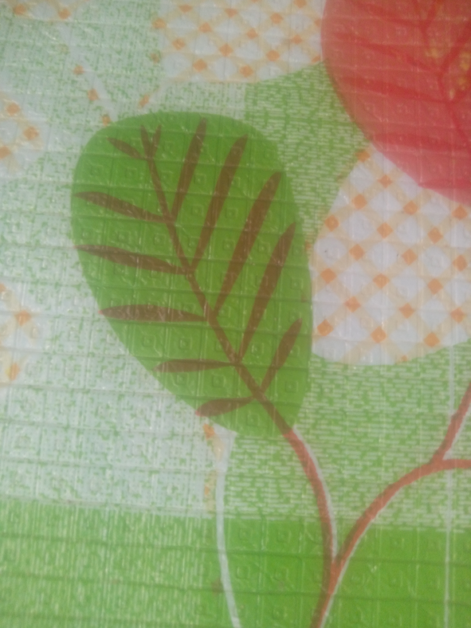 The image is a close-up, slightly blurry shot of a patterned surface, likely a mat or fabric, with a floral or fruit motif. The dominant colors are various shades of green, with accents of red, orange, and white.
The main subject in the foreground is a large, green leaf with prominent brown veins. The leaf has a textured surface, appearing to be made of small, repeating square patterns. Behind the leaf, a large, red, circular shape is visible, possibly representing a fruit like an apple or tomato, although it is mostly cropped out. There are also lighter, patterned areas with a grid of small orange squares on a white background, interspersed with the green.
The background consists of a lighter green area with a very fine, almost pixelated texture. Thin, reddish-brown lines, resembling stems or branches, weave through the composition, connecting to the leaf and extending out of frame.
There is no discernible activity or event taking place, as the image is a static depiction of a pattern. The time of day, weather, and emotions are not applicable as there are no living subjects or environmental elements. No text is visible in the image. The overall impression is that of a decorative surface.