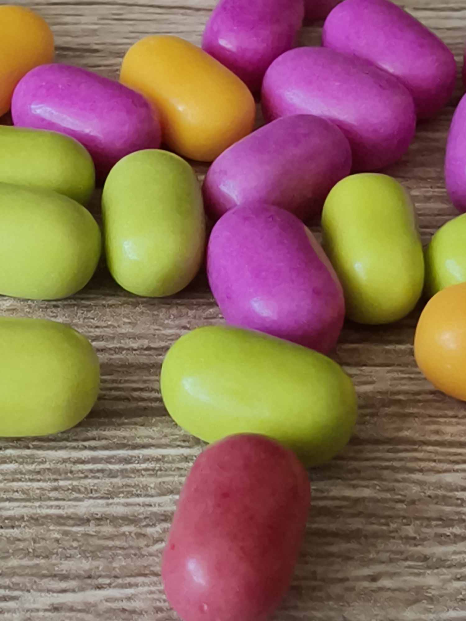 A close-up shows a collection of candy-coated, pill-shaped sweets scattered on a textured surface. The candies are in various colors, including pink, green, orange, and red. The lighting appears soft, reflecting off the smooth surfaces of the candies. The background has a horizontal, wood-like grain. There is no information available to suggest that the image was taken in Kryvyi Rih, Ukraine. The location cannot be determined from the image. No people are visible.