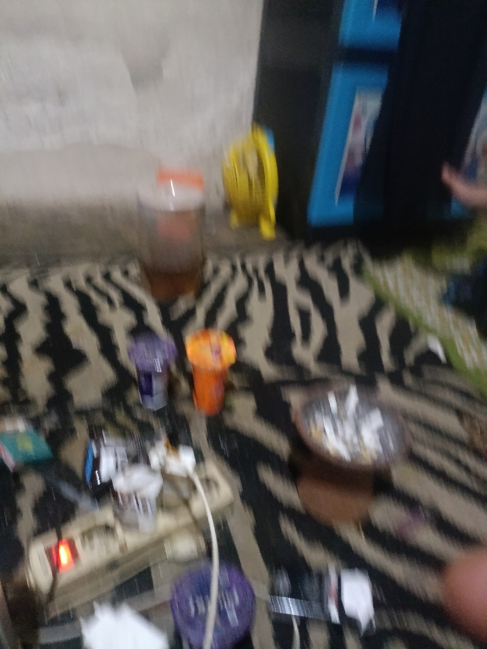The image shows a cluttered surface covered with a patterned rug with black and white stripes. The rug is surrounded by a white wall. A power strip with multiple plugs is located in the center of the image. On the right side of the image, there is a partially visible person's hand. There is a partially visible yellow object, possibly a toy. There are several plastic containers, some with their lids off, scattered across the surface. The scene is dimly lit, with a soft light emanating from an unknown source.