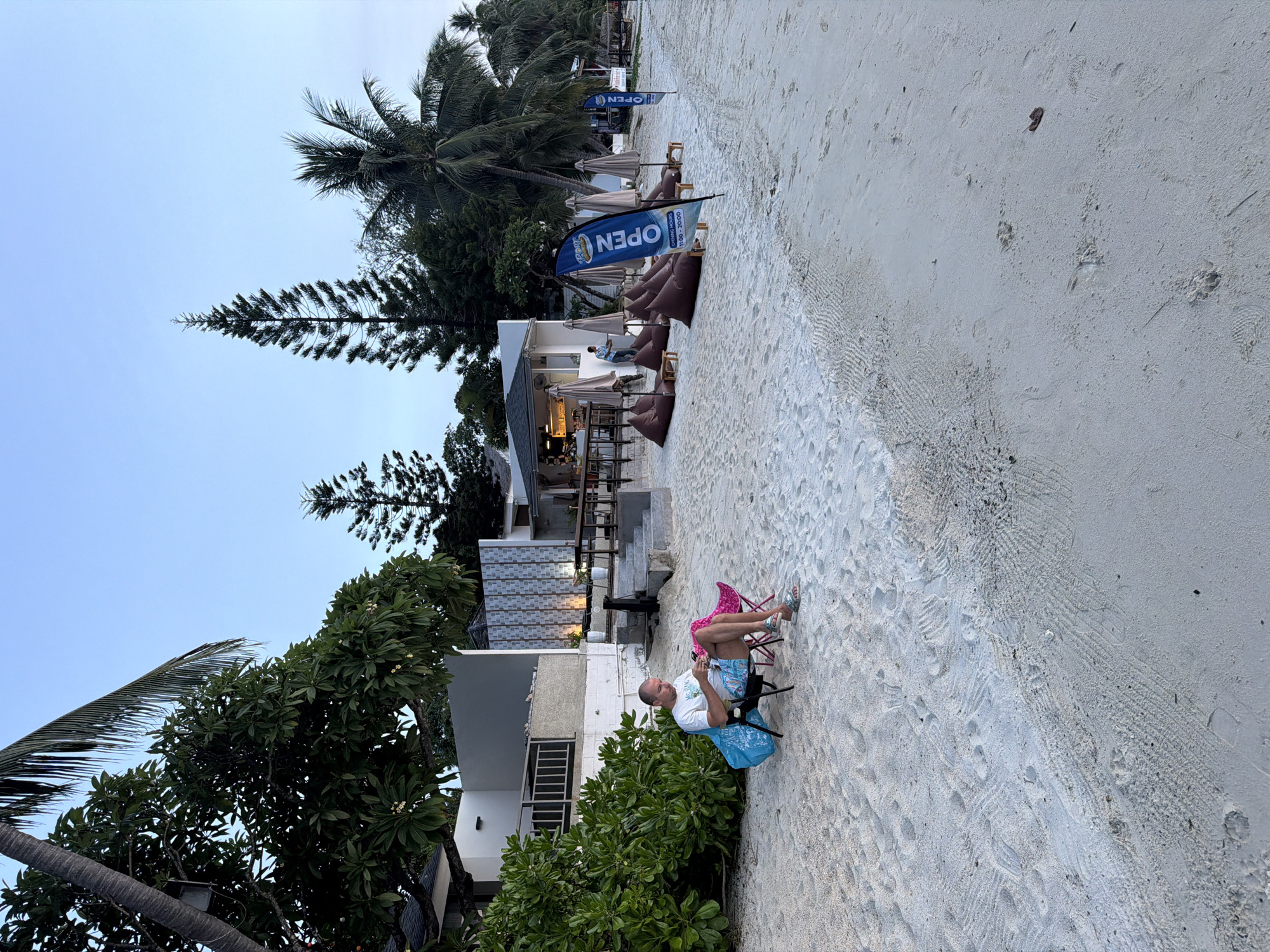 The image depicts a beach scene at dusk in Baan Bang Rak, Thailand. A bald, middle-aged man is seated in a portable chair on the sandy beach, looking at his phone. He is wearing a white t-shirt and patterned shorts. A blue tote bag is placed beside his chair. In the background, a low-rise building with an outdoor seating area appears to be a restaurant or cafe, with tables and chairs set up on a raised platform. Several large beanbag chairs are arranged on the sand in front of the establishment, along with closed umbrellas. Two tall, narrow flags with the word "OPEN" are visible. Palm trees and other foliage surround the building and extend along the coastline. The sky is a pale blue, indicating the transition from day to night. The beach is covered in light-colored sand, with visible footprints and tire tracks.