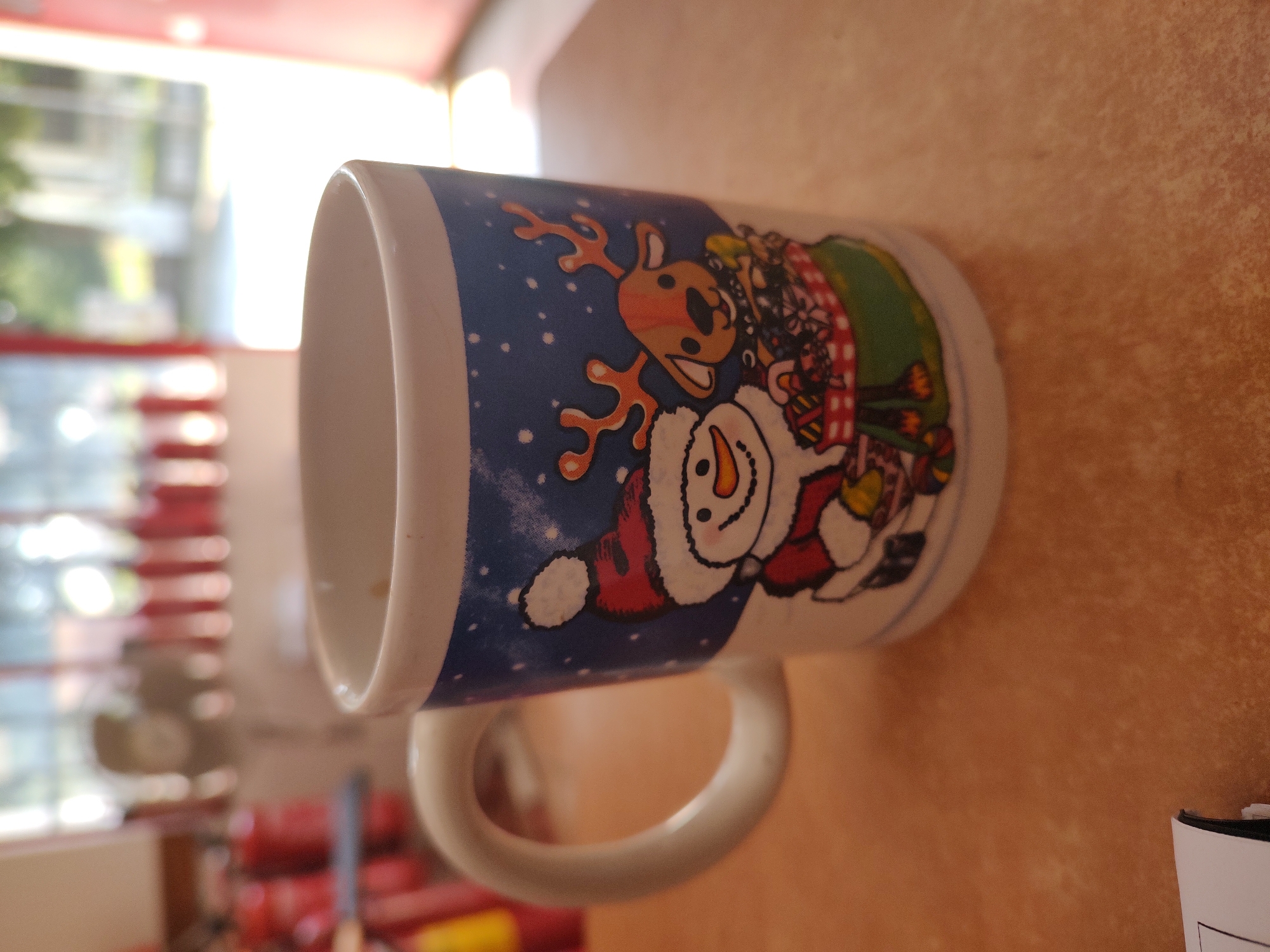 A close-up, eye-level shot shows a white ceramic mug decorated with a Christmas theme, featuring a snowman wearing a red and white Santa hat and a brown reindeer with antlers, both standing in front of a green basket filled with gifts. The mug has a white handle and a blue background with white dots, resembling snow. The mug is placed on a textured, orange-brown surface, likely a table. In the blurred background, there are red fire extinguishers and vertical red elements behind a window, suggesting an indoor setting, possibly a workshop or storage area. The overall lighting suggests daytime, with light streaming in from the background. The image composition focuses on the mug, with the background out of focus, indicating it is not the primary subject. No specific location in Tervel, Bulgaria can be determined from the visual information provided.
