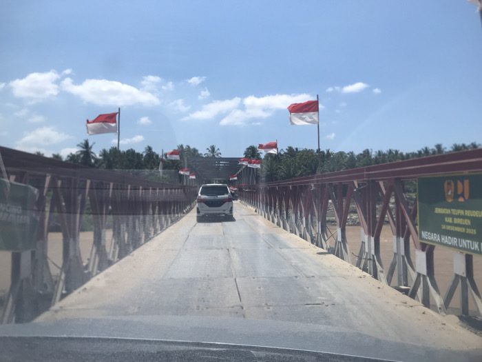 This image captures a daytime scene on the Teupin Reudeuep Bridge in Ceubrek, Bireuen Regency, Indonesia. A white SUV is observed driving away from the viewer on the concrete or asphalt surface of the bridge, which features a red metal truss structure. A wide, muddy brown river flows beneath the bridge. Multiple red and white Indonesian flags are prominently displayed along the bridge's framework and on poles. The sky above is clear blue with scattered white clouds. On the right side of the bridge, a green sign with white text is visible, stating "JEMBATAN TEUPIN REUDEUEP KAB. BIREUEN 18 DESEMBER 2025 NEGARA HADIR UNTUK..." (Teupin Reudeuep Bridge, Bireuen Regency, 18 December 2025, The State is Present For...). Lush green vegetation, including numerous palm trees, lines the riverbanks in the background. The photograph is taken from inside another vehicle, evidenced by a slight dashboard reflection at the bottom edge.