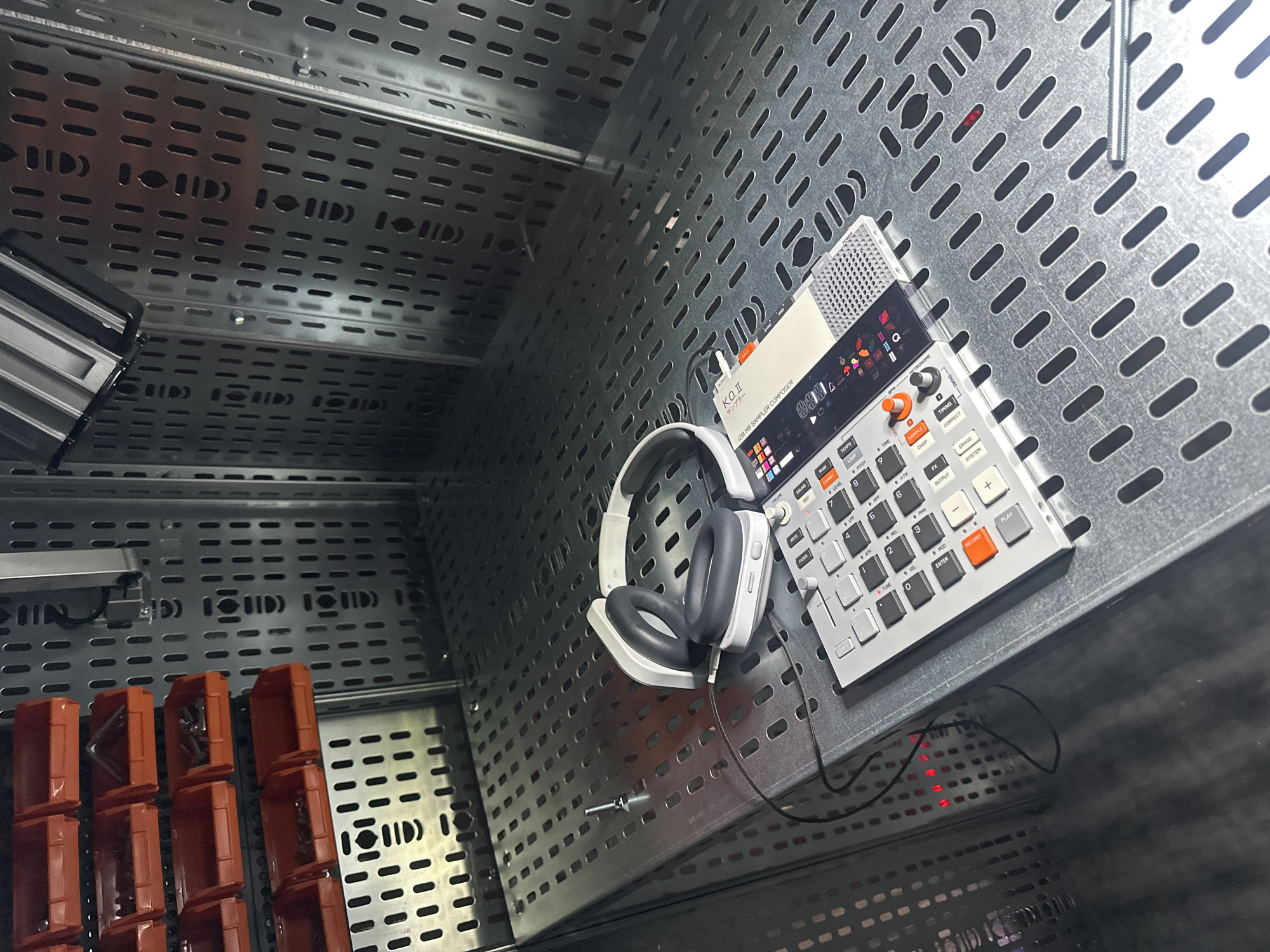 The image captures a close-up of music production equipment set within an industrial or technical environment, likely a studio or workshop in London, United Kingdom. The main subjects are a white and grey electronic sampler/composer and a pair of white over-ear headphones.
The sampler, identified as a Teenage Engineering PO-33 K.O. II, is positioned on a metallic, perforated surface resembling a cable tray or a modular shelving unit. The device features a compact design with a small screen displaying graphical elements, numerous grey and black buttons, orange knobs, and a white fader. Prominently visible text on the device includes "KO II," "サンプラー" (Sampler), "128 MB SAMPLER COMPOSER," "TEMPO," "SAMPLE," "CHOP," "PLAY," "RECORD," "ERASE," and controls for various audio parameters such as "PITCH," "PAN," "LEVEL," "FX," "ATK," and "REL."
A pair of white headphones with dark grey earcups is placed next to the sampler, connected by a black cable. The metallic perforated surface extends across the background, characterized by a uniform grid of elongated holes. In the lower-left, several orange modular containers or trays are visible, suggesting storage for tools or components. A silver screw or bolt lies on the perforated surface near the headphones. Beneath the sampler, subtle red indicator lights can be seen glowing through the perforations, hinting at other electronics or power sources below. The overall scene suggests a setup for creative audio work or music production, ready for immediate use.