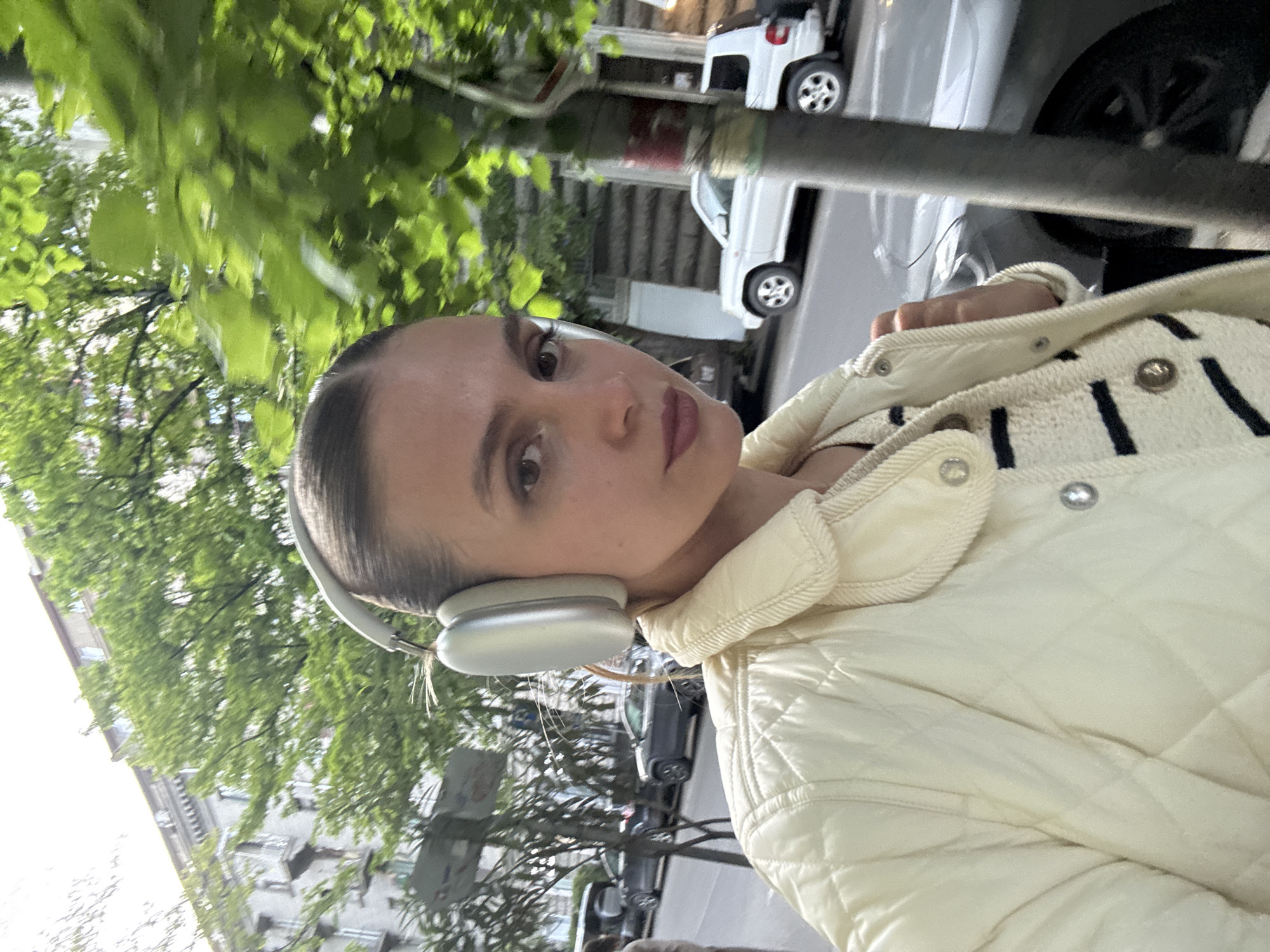 An outdoor, slightly rotated portrait captures a young adult female on a city street, likely in Tbilisi, Georgia. The subject, positioned centrally, looks directly at the camera with a neutral expression. She has dark hair pulled back and is wearing light makeup, including dark eyeliner and a berry-toned lip color. She wears silver over-ear headphones, a light cream or yellow quilted jacket, and a striped garment underneath.
The setting appears to be a vibrant urban or residential area during daytime. Lush green trees with full foliage dominate the background and upper left, suggesting spring or summer weather. Several vehicles are visible on the street behind her, including a prominent white car and a small white toy car or scooter on what appears to be a sidewalk. Buildings are partially visible through the trees and above the parked cars. The lighting is bright, indicating good weather. There is no visible text in the image. The overall activity seems to be a casual moment captured outdoors, possibly a selfie taken while walking or pausing.