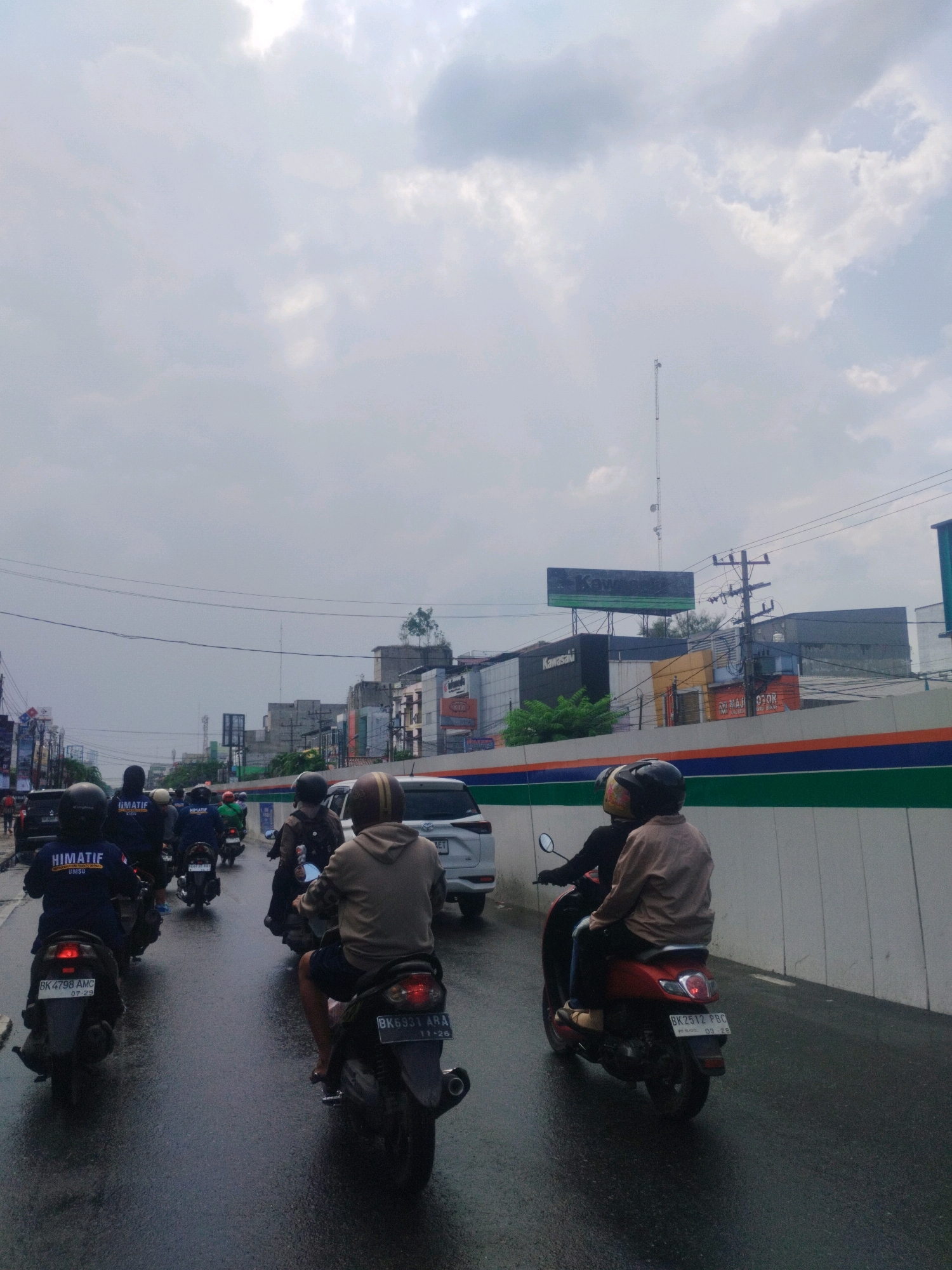 The image depicts a street scene in the city of Medan, Indonesia, on an overcast day. Several motorcycles are on a wet asphalt road, indicating recent rain or damp conditions. In the foreground, two individuals are riding a red scooter, with the passenger wearing a light brown jacket and the driver in dark clothing. To the left, a line of motorcycles is moving away from the camera. The riders on these motorcycles are wearing dark jackets with the text "HIMATIF" and "UMSU" visible on the back, suggesting they are part of a student organization. A white car is visible behind the motorcycles. In the background, buildings line the street, some featuring signage such as "Kawasaki". Overhead, power lines crisscross the sky, and a communication tower can be seen in the distance. The overall atmosphere is one of urban traffic under a cloudy sky.